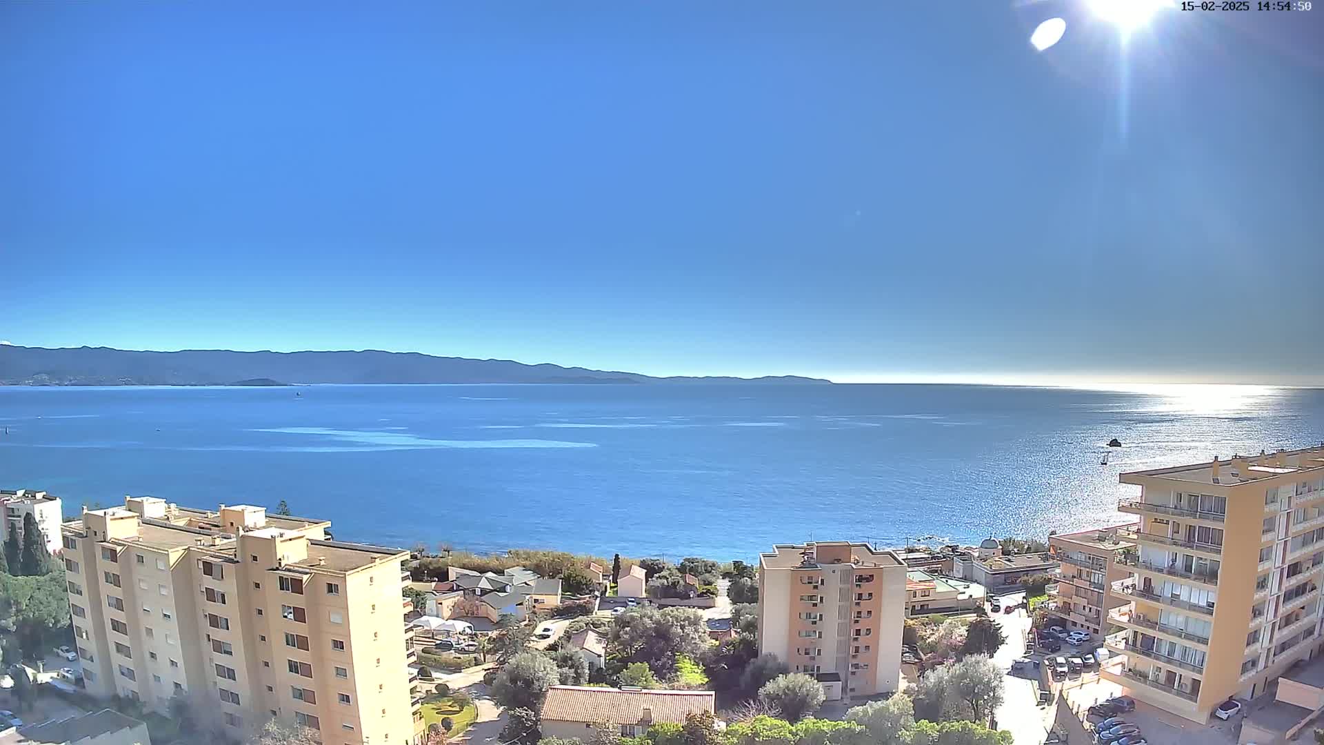 A sunny day reveals a coastal town nestled between a calm, blue sea and distant mountains.
