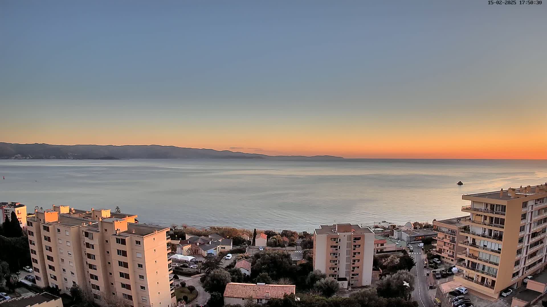 A coastal sunset scene shows calm water, distant mountains, and several apartment buildings overlooking the sea.