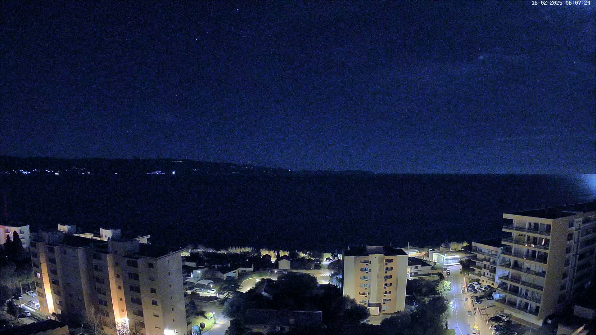 A nighttime aerial view shows a dark body of water, a coastline with several apartment buildings, and a clear, starry sky.