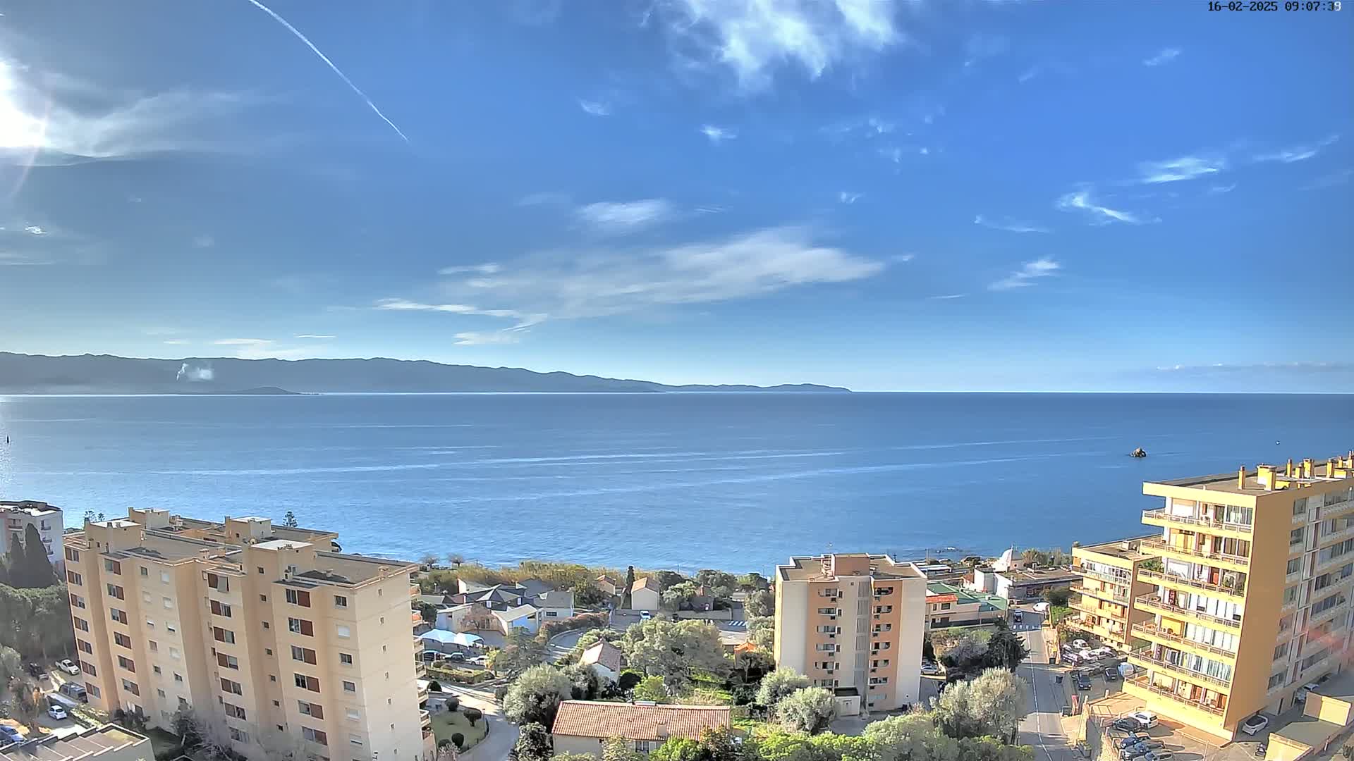 A mostly sunny day overlooks a calm blue sea with distant mountains and several apartment buildings situated near the water's edge.
