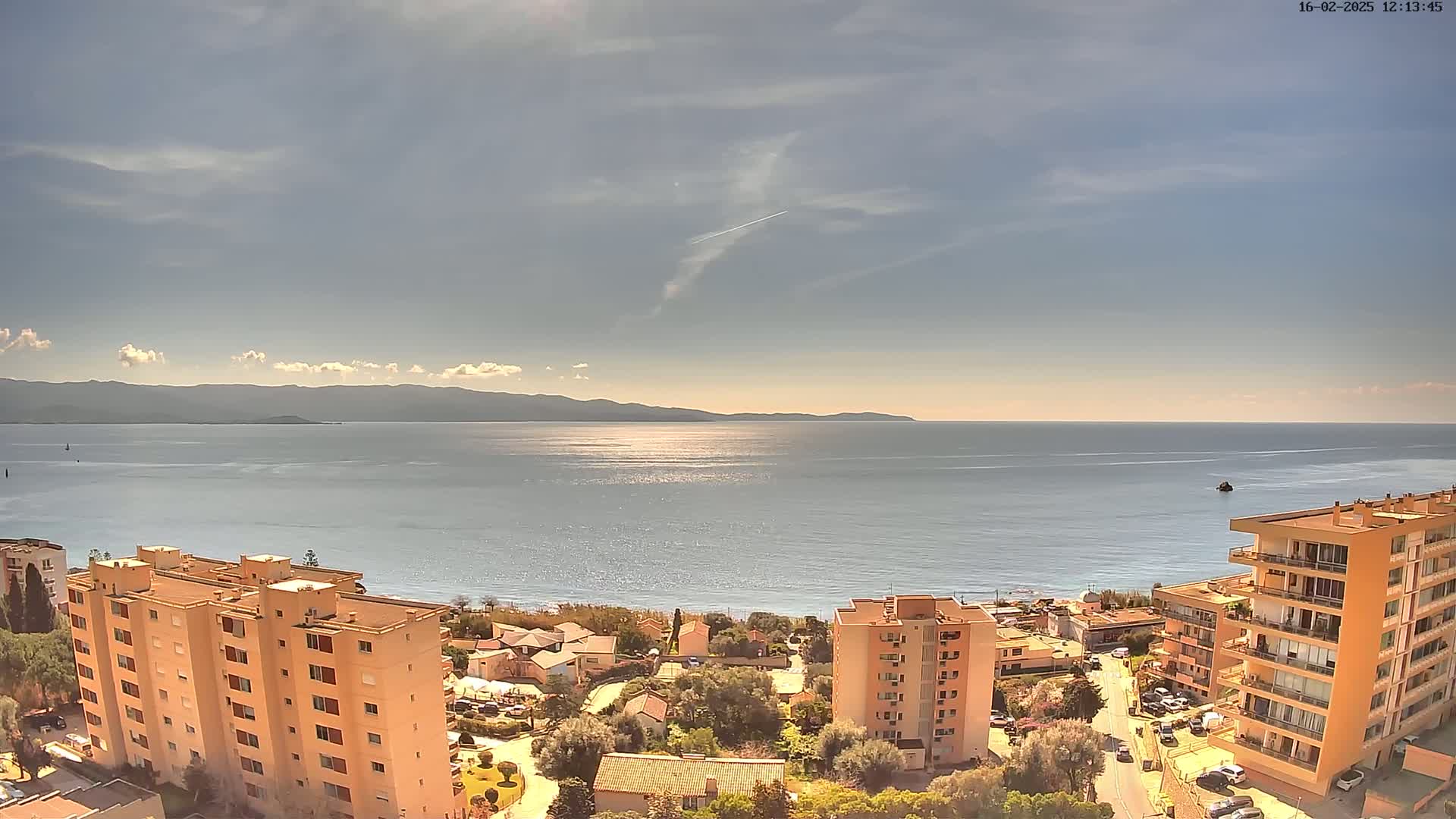 A sunny day reveals a calm sea stretching to distant mountains, with several apartment buildings and a small town nestled along the coast.