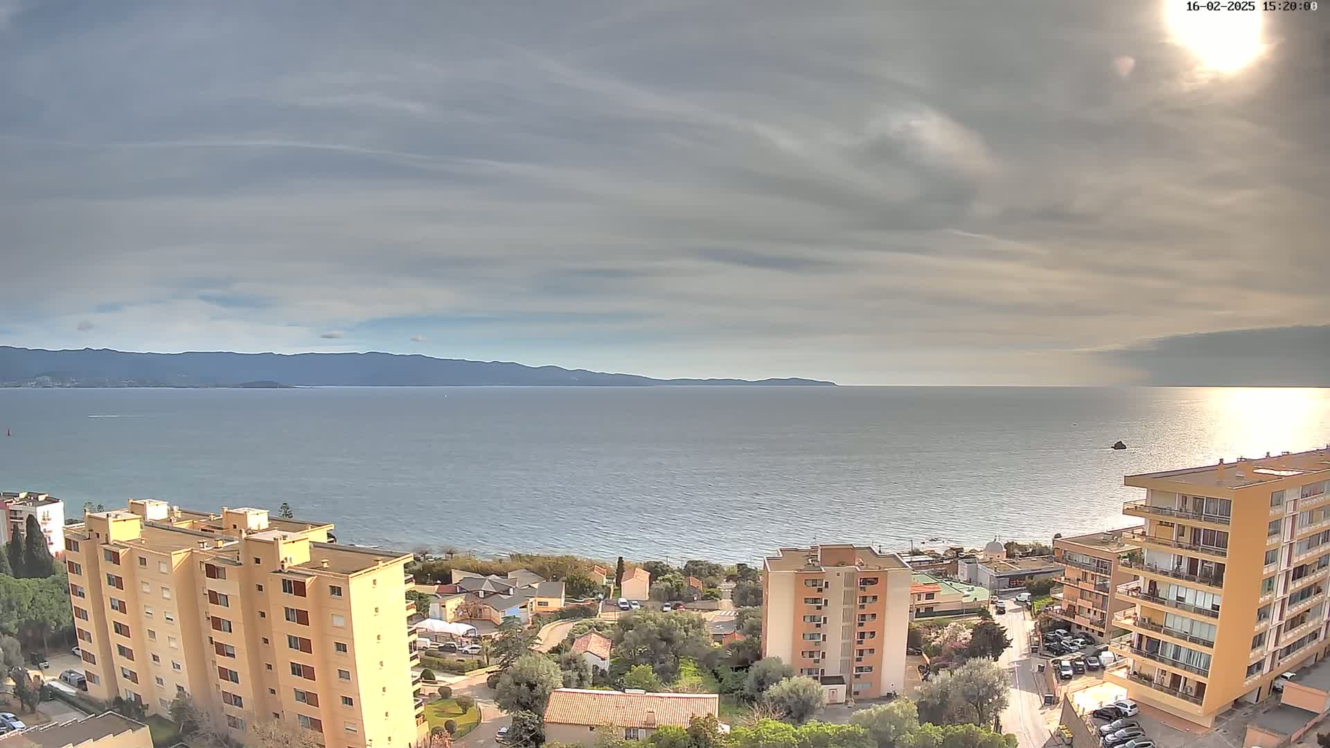 A coastal town with several apartment buildings overlooks a calm sea under a mostly cloudy sky, with mountains visible in the distance.