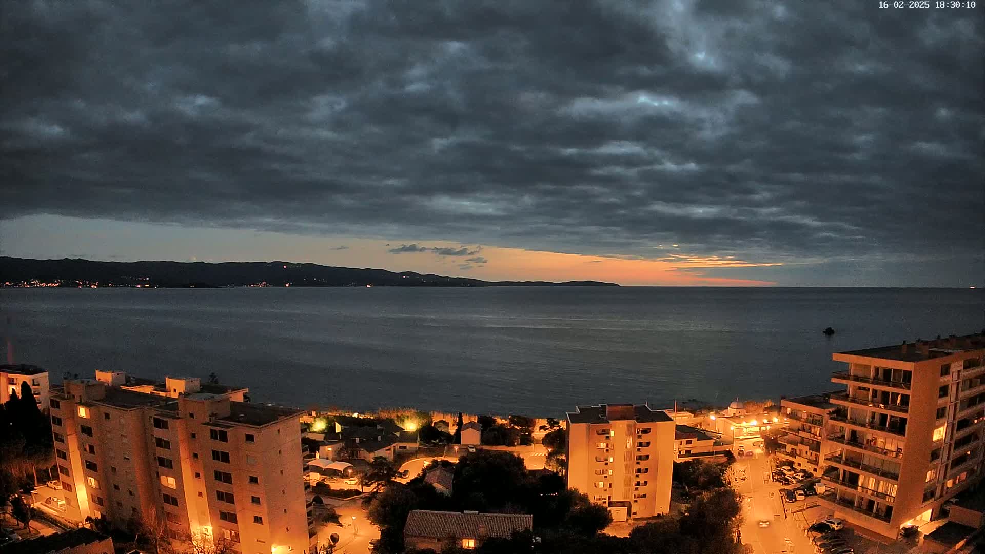 A nighttime view of a coastal town with apartment buildings illuminated under a dark, cloudy sky with a hint of orange sunset on the horizon over a calm sea.