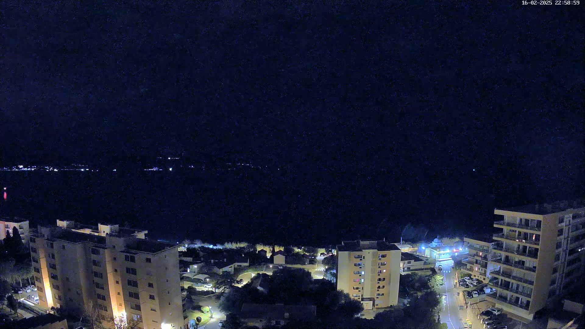 A nighttime view of a coastal town with several multi-story buildings illuminated against a dark, clear sky over calm water.