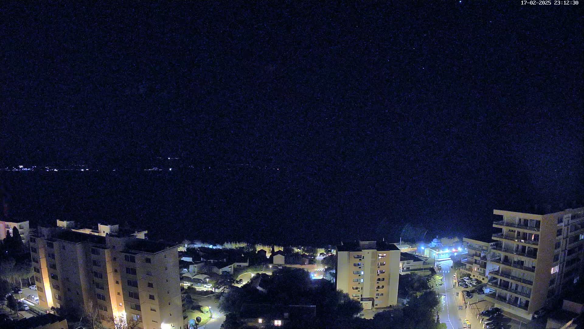 A nighttime view of a coastal town shows several apartment buildings illuminated against a dark, star-filled sky.