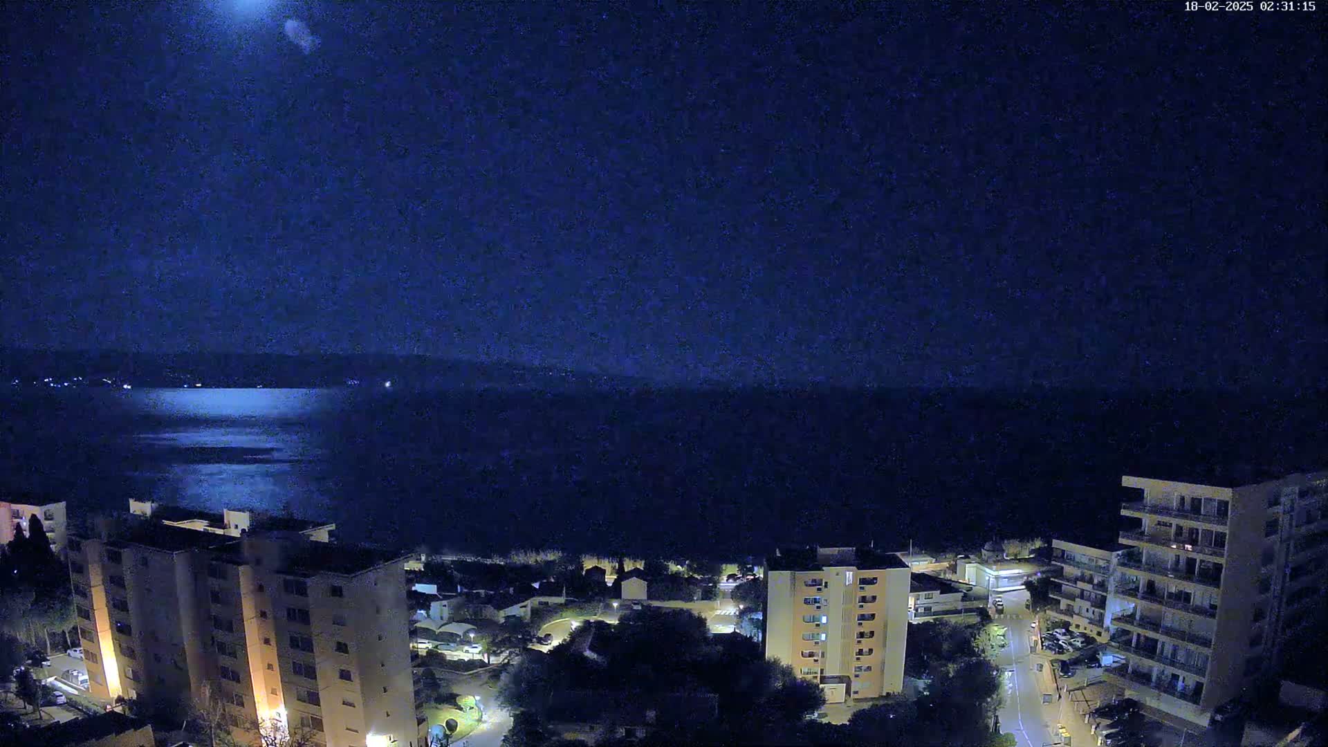A nighttime view of a calm body of water with a distant shoreline, overlooked by several multi-story buildings with lights on, under a dark, clear sky.