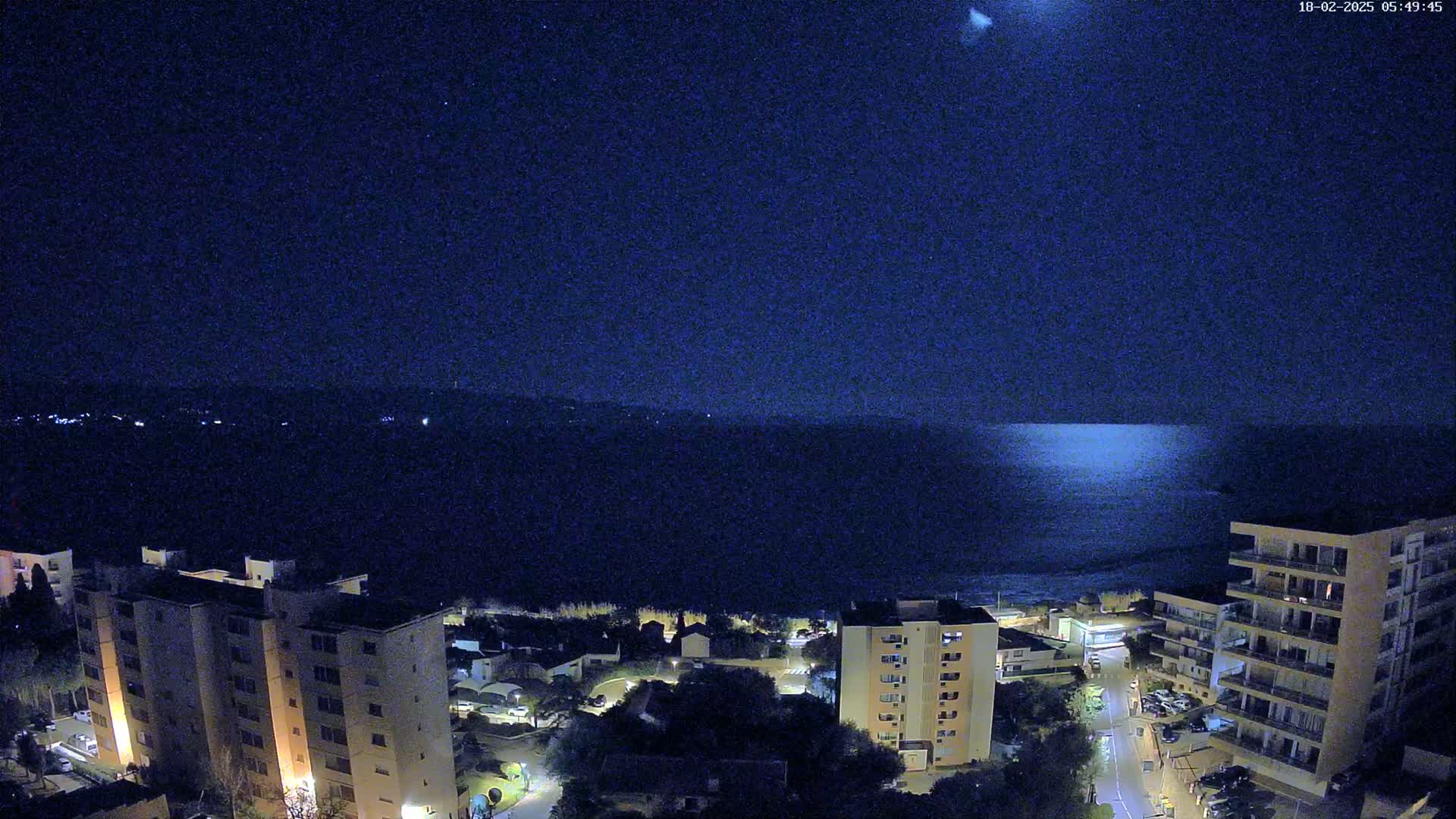 A nighttime view shows a calm sea reflecting light under a dark, clear sky, with illuminated apartment buildings and surrounding streets below.