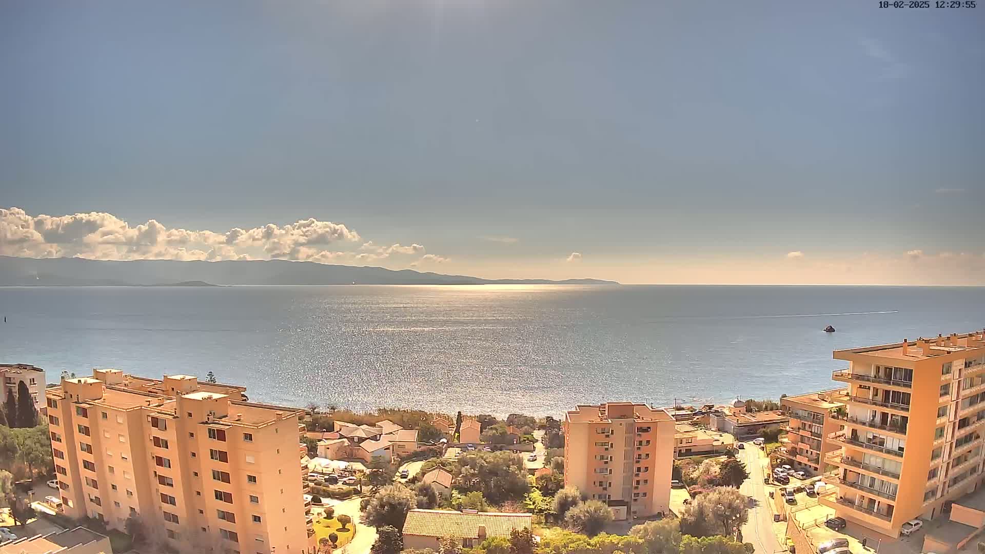 A sunny day reveals a coastal town with several multi-story buildings overlooking a calm sea and distant mountains under a partly cloudy sky.