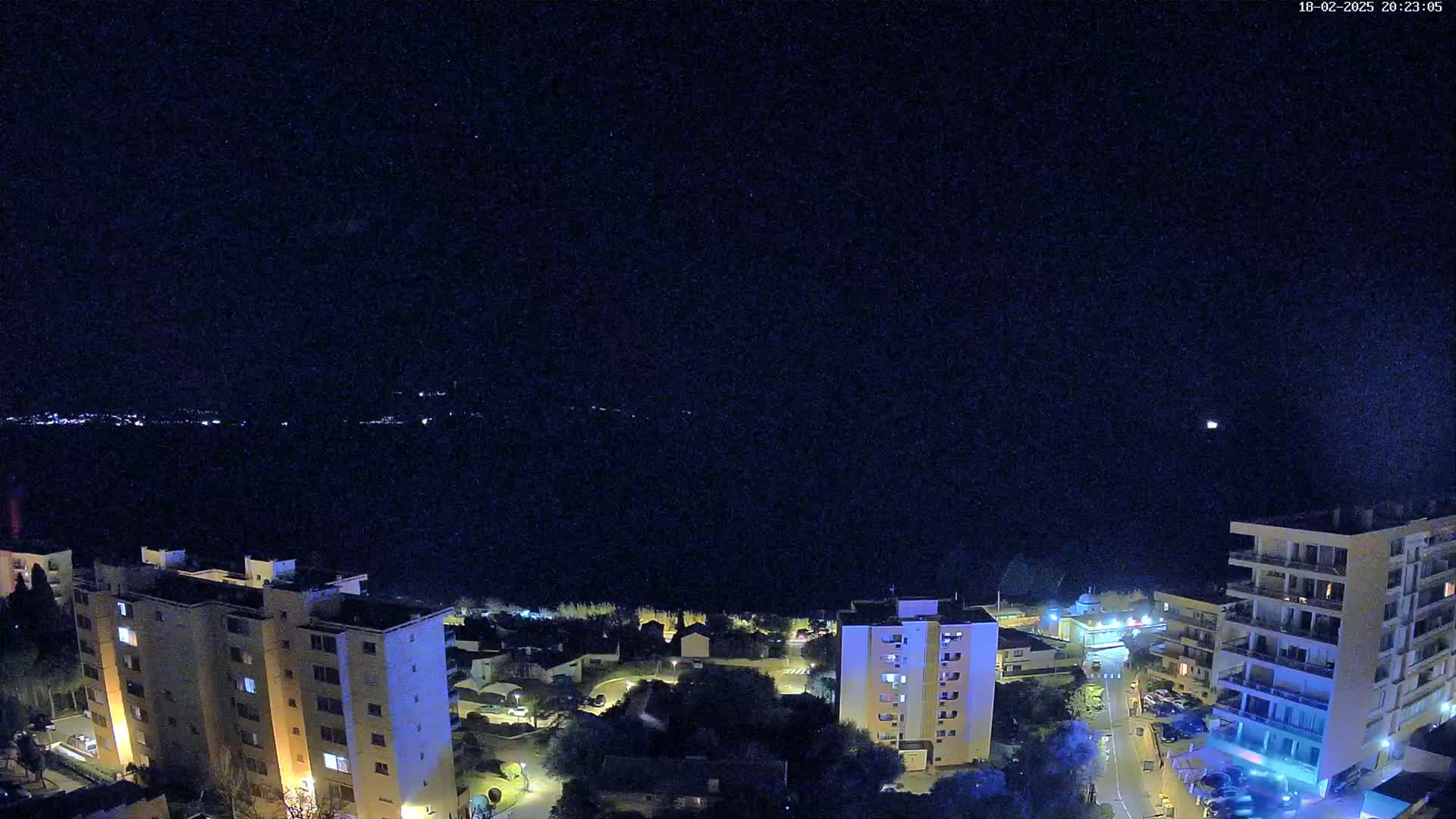A nighttime, aerial view shows a coastal town with apartment buildings illuminated against a dark sky dotted with stars, overlooking a dark body of water.