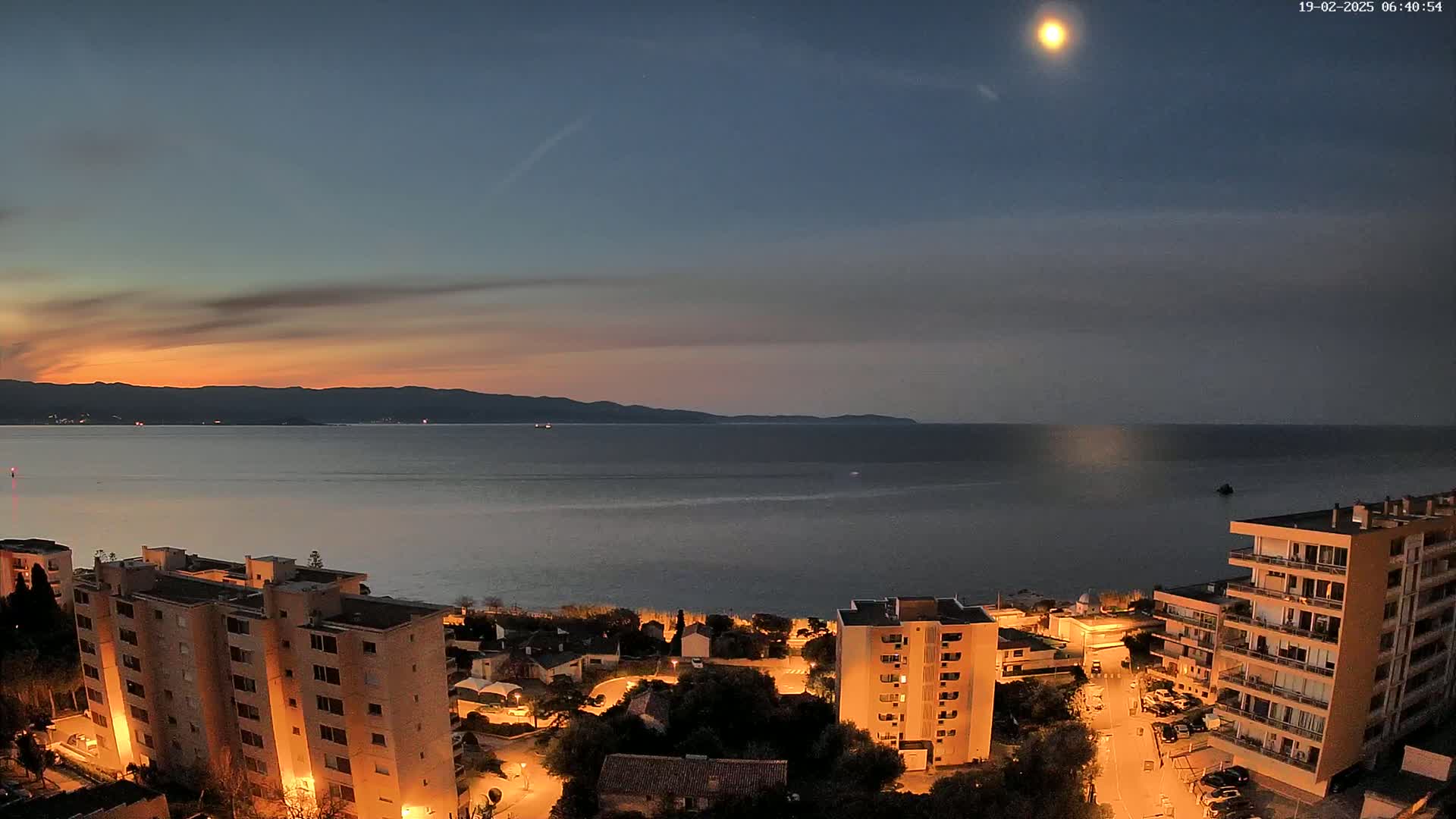 An aerial, nighttime view shows a calm body of water with mountains in the distance under a twilight sky with a bright moon.