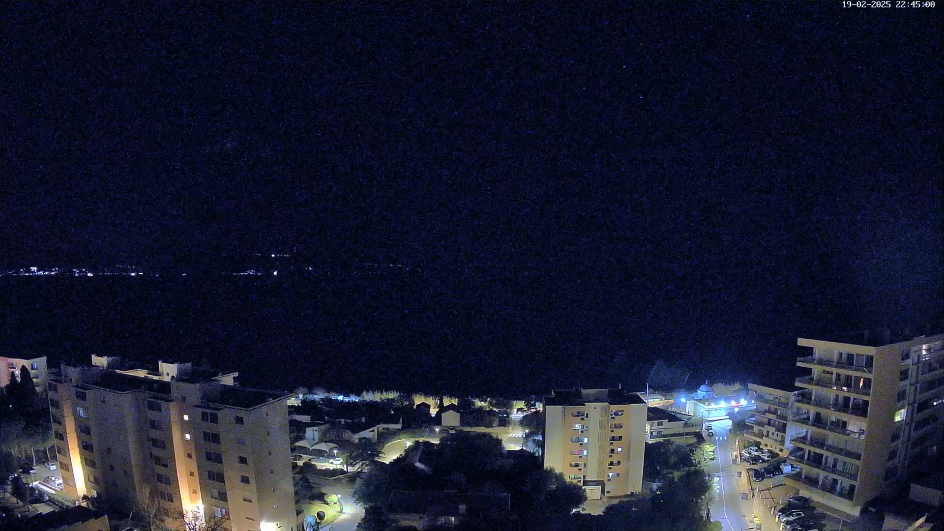 A nighttime aerial view shows a coastal town with several multi-story buildings illuminated against a dark, clear sky.