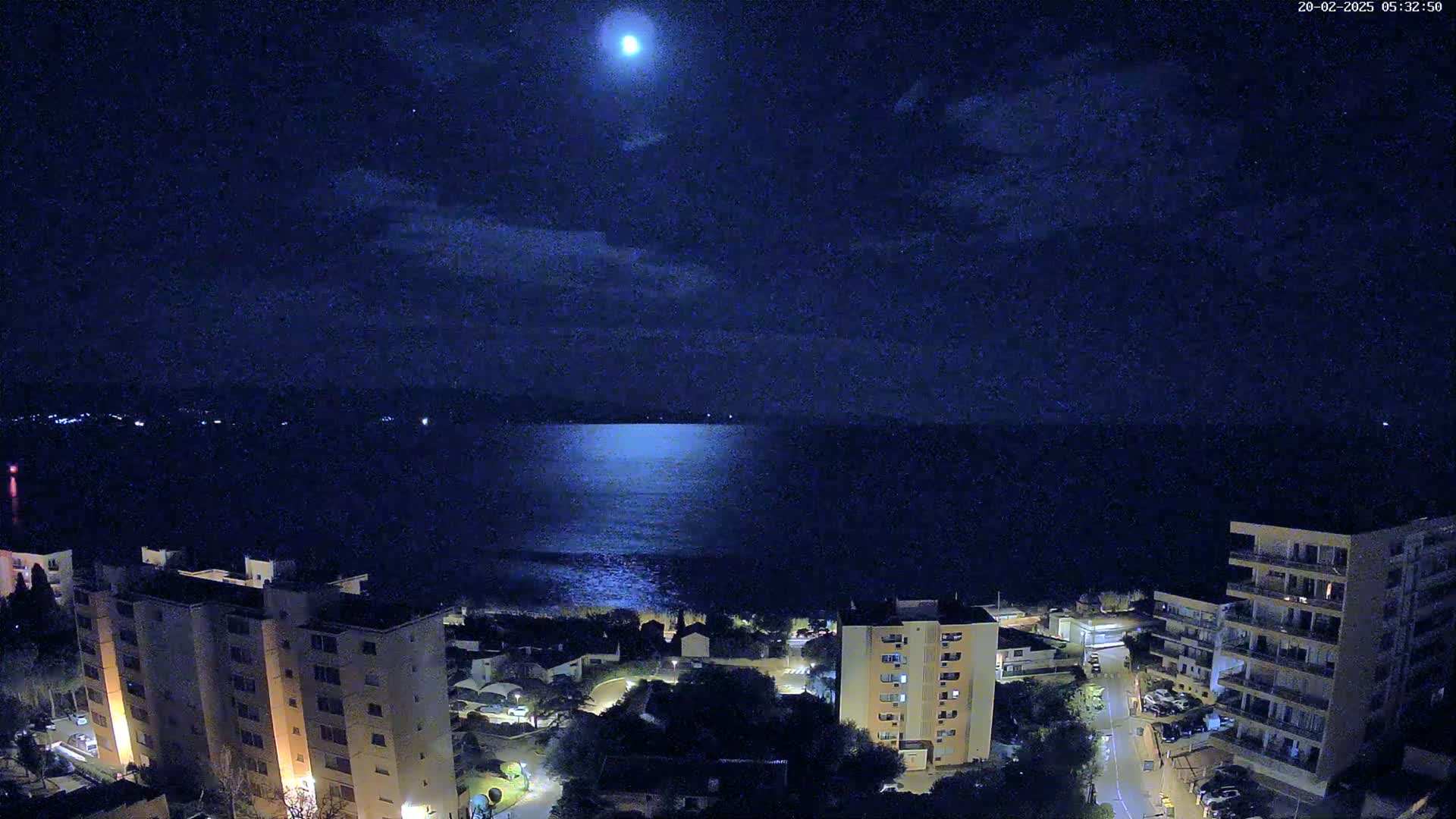 A bright moon shines over a calm, dark body of water at night, illuminating a coastal town with apartment buildings below.