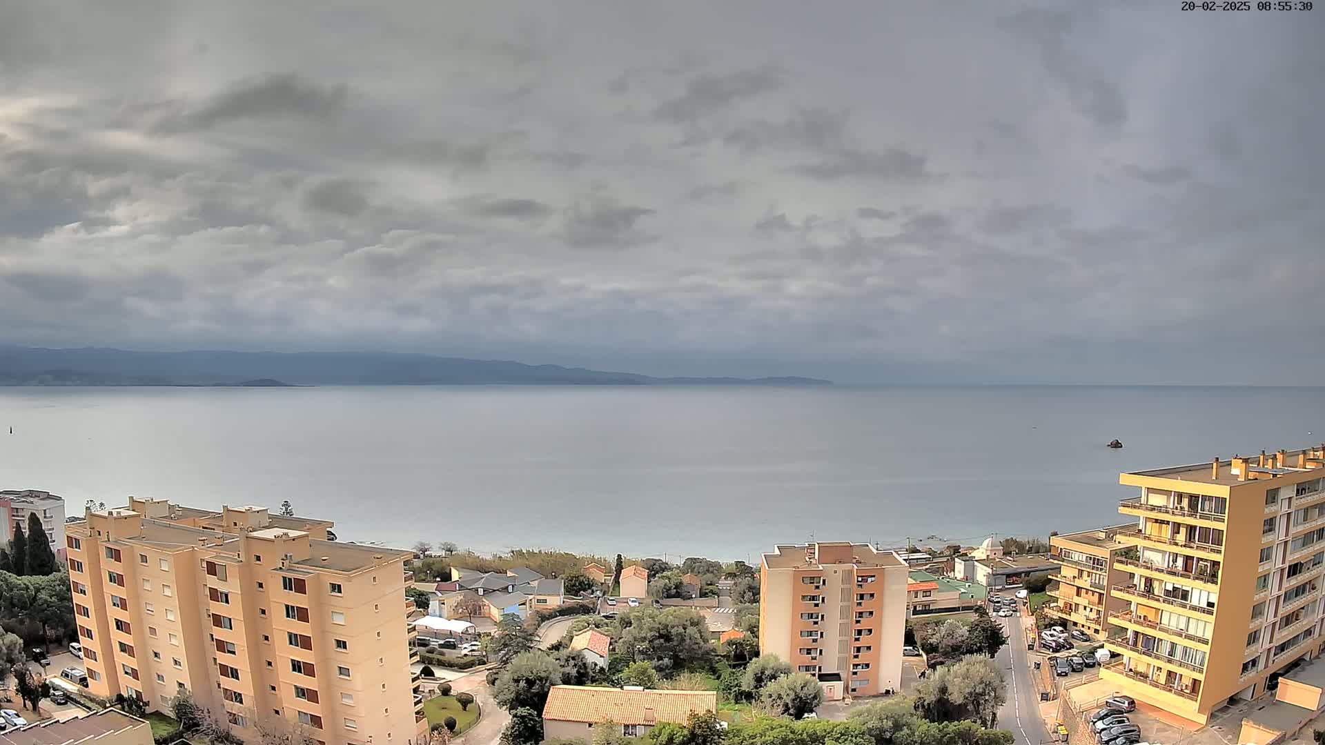 An overcast sky overlooks a calm sea with a coastline of buildings and trees.