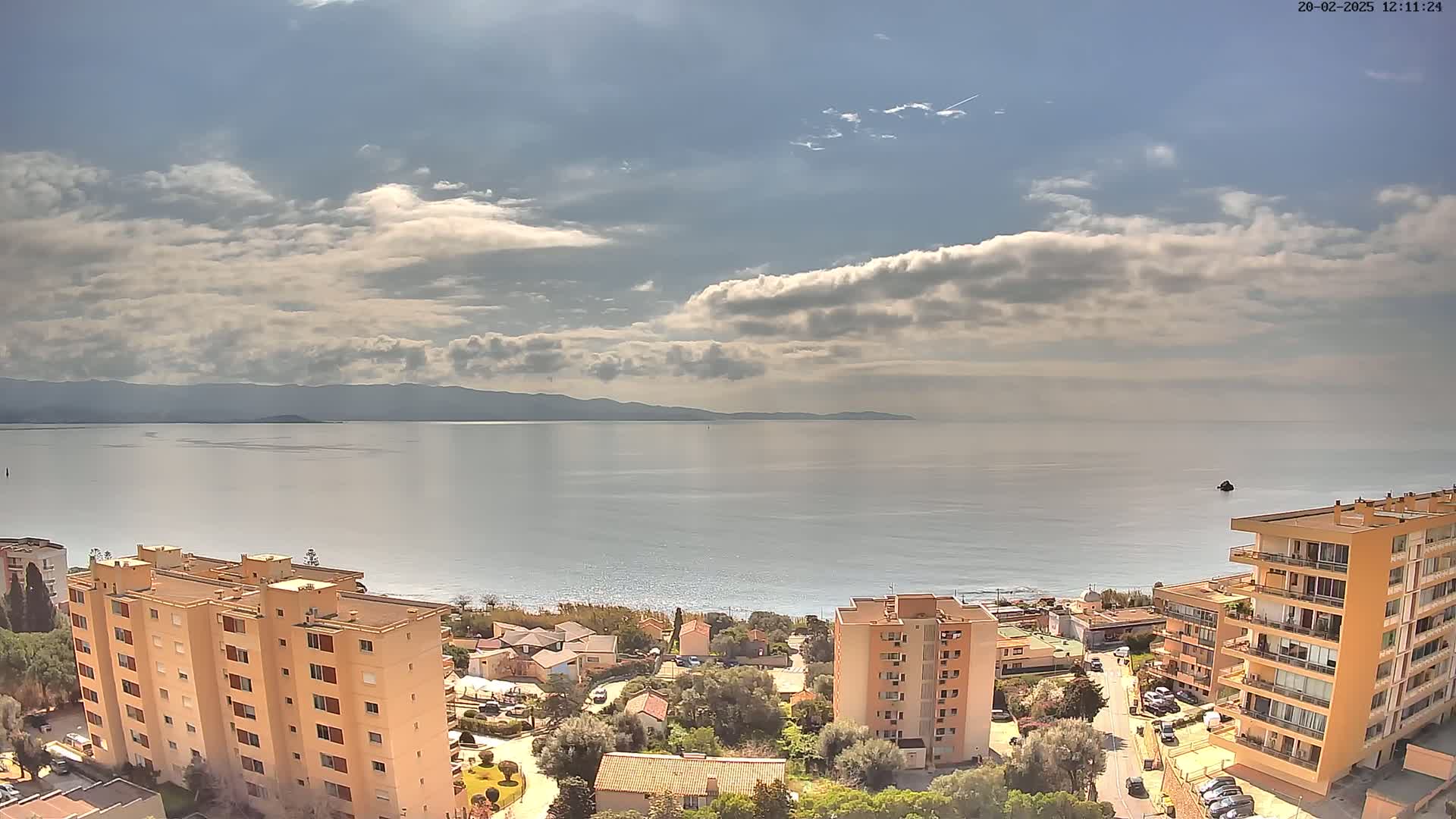 A partly cloudy day reveals a coastal scene with calm water, distant mountains, and several multi-story buildings situated near a beach.