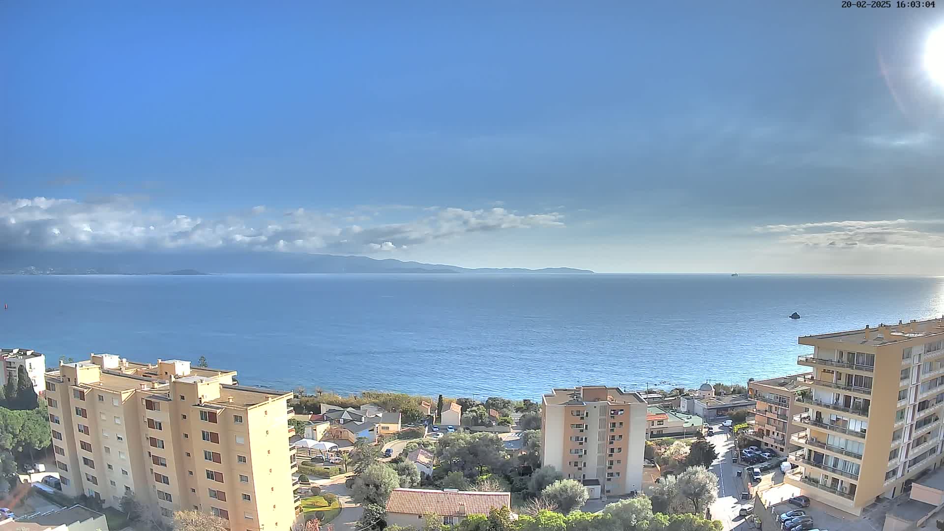 A coastal town with apartment buildings overlooks a calm blue sea under a mostly sunny sky with a few scattered clouds and distant mountains.