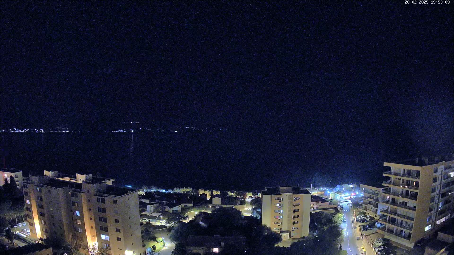 A nighttime view overlooking a coastal town with apartment buildings, streets, and a dark body of water in the distance under a clear, starless sky.