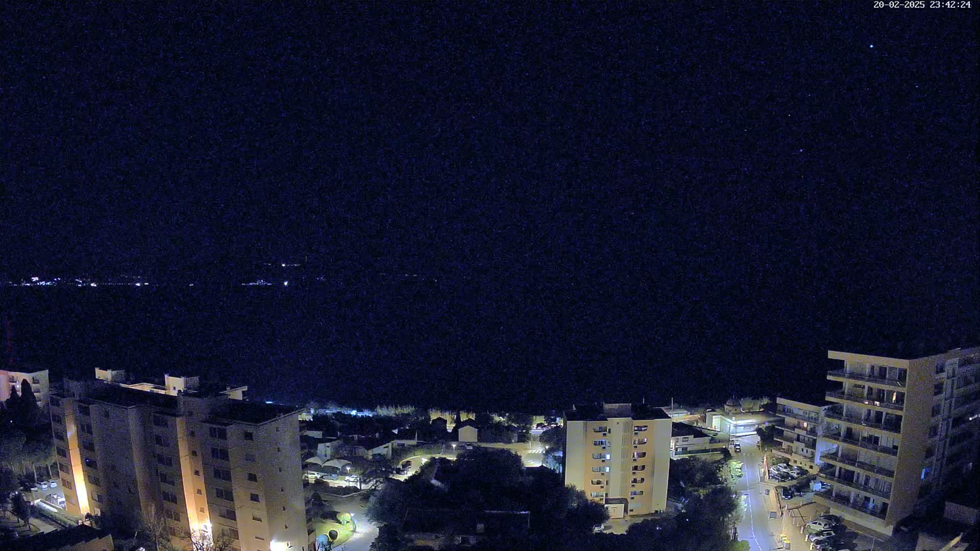 A nighttime aerial view shows a coastal town with several multi-story buildings under a clear, starry sky.