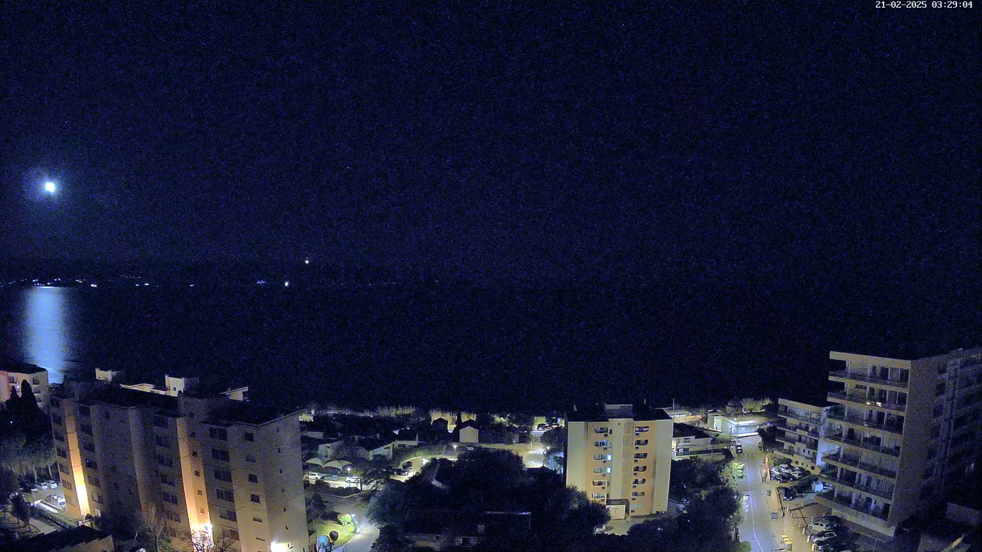 A nighttime view of a calm body of water reflecting a bright moon, with illuminated buildings and trees lining the shore.