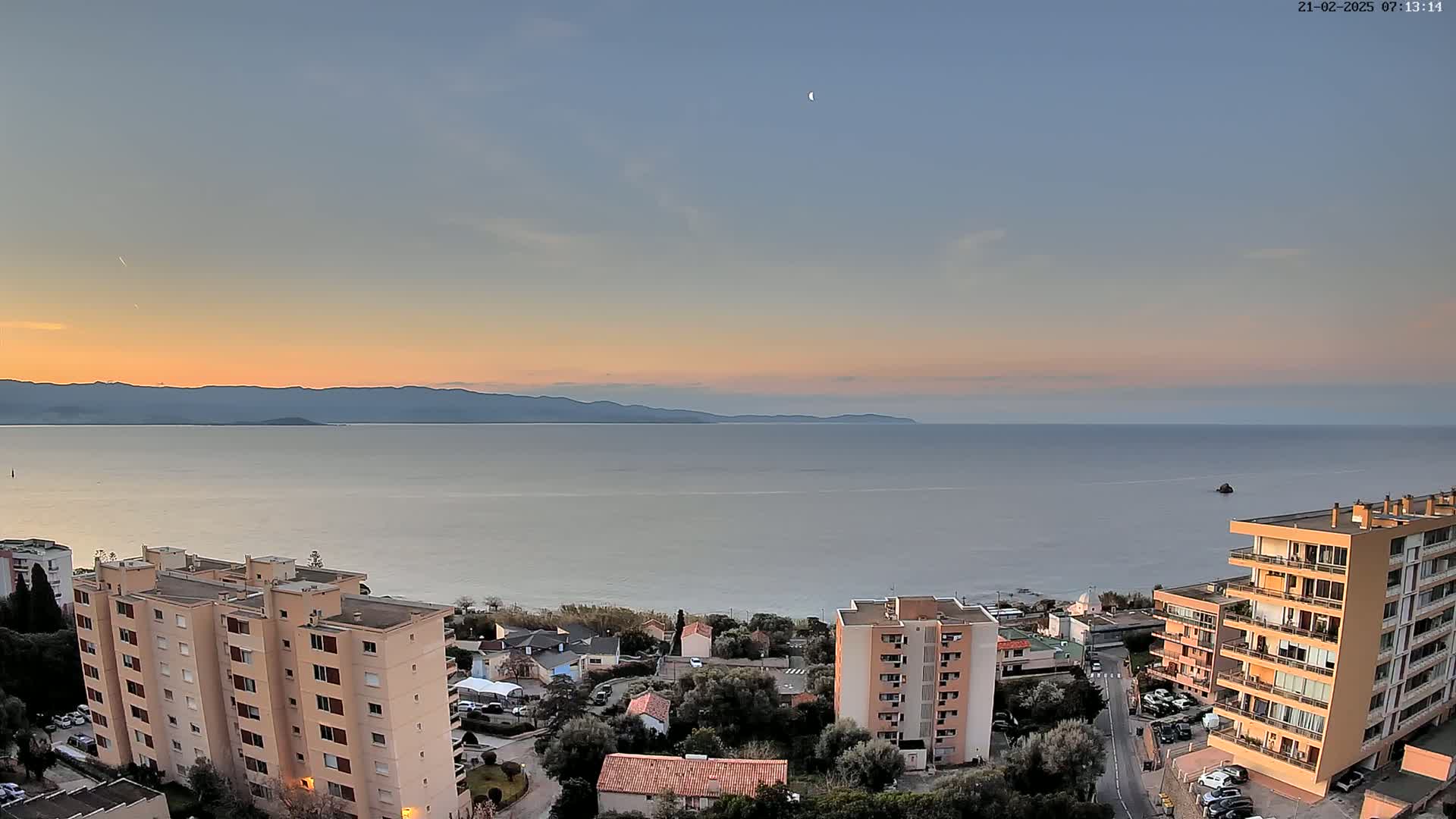 A calm sea stretches to distant mountains under a pale pink and blue sunset sky, with a crescent moon visible, and several apartment buildings situated on a hillside overlooking the water.