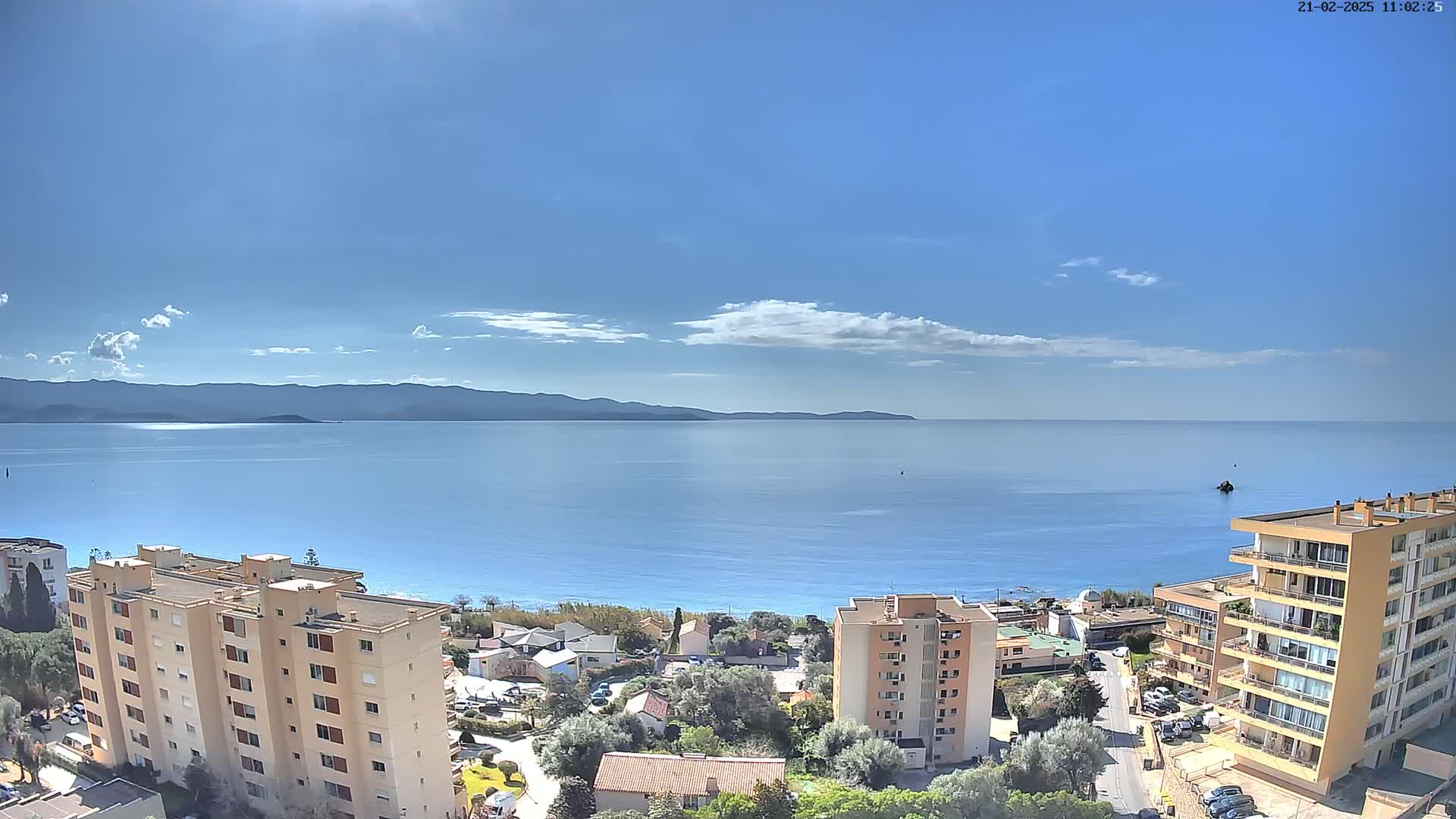 A sunny day overlooking a calm blue sea with distant mountains, and apartment buildings in the foreground.