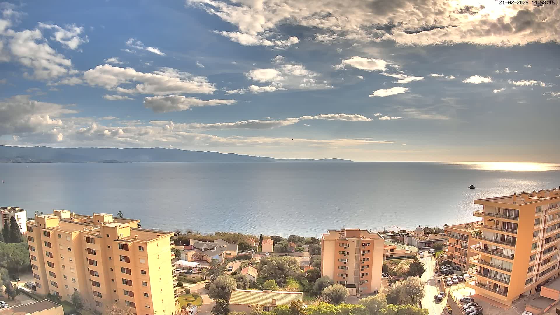A partly cloudy day overlooks a calm sea with distant mountains, buildings, and trees in the foreground.