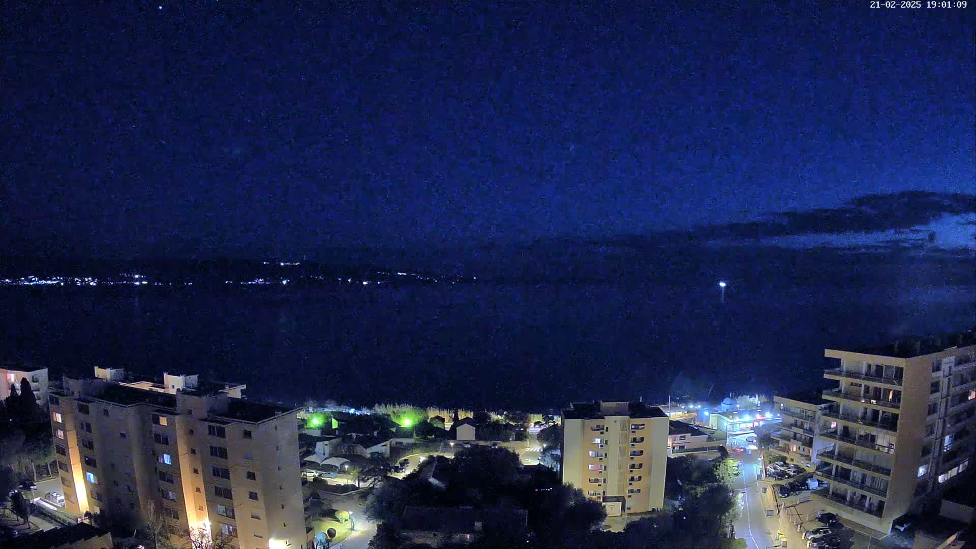 A nighttime, high-angle view shows a dark body of water with a distant city skyline illuminated, overlooked by several multi-story apartment buildings under a mostly clear, dark blue sky.