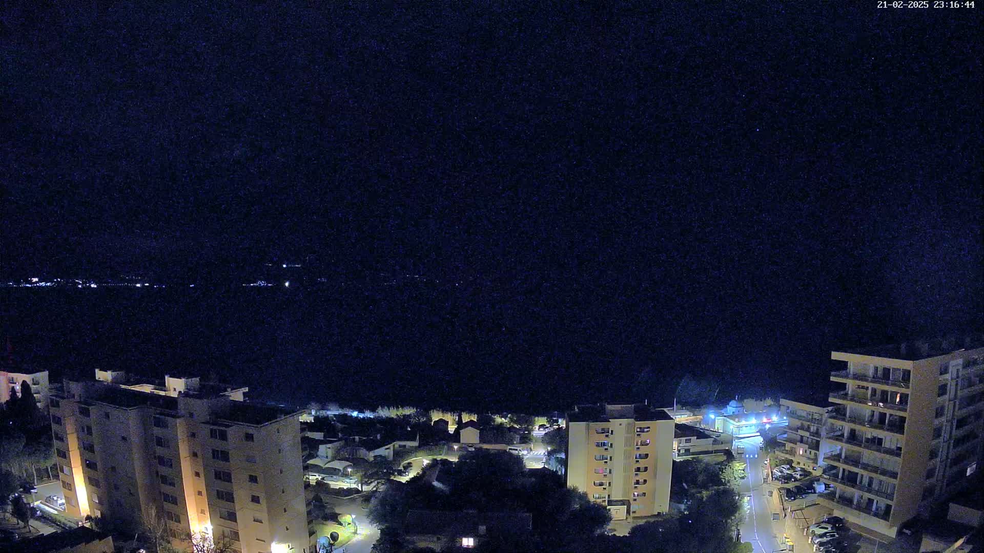 A nighttime, aerial view of a coastal town shows several multi-story buildings illuminated against a dark, starlit sky over a calm body of water.