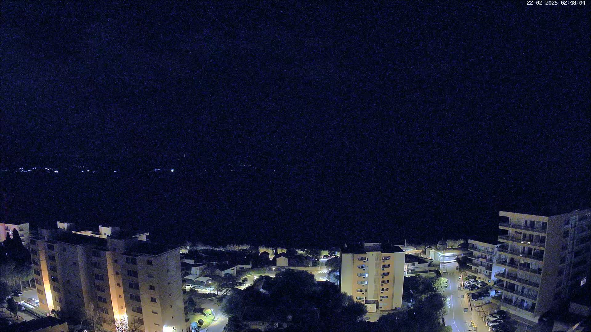 A nighttime aerial view of apartment buildings and a street, with a dark sky and distant lights across a body of water.