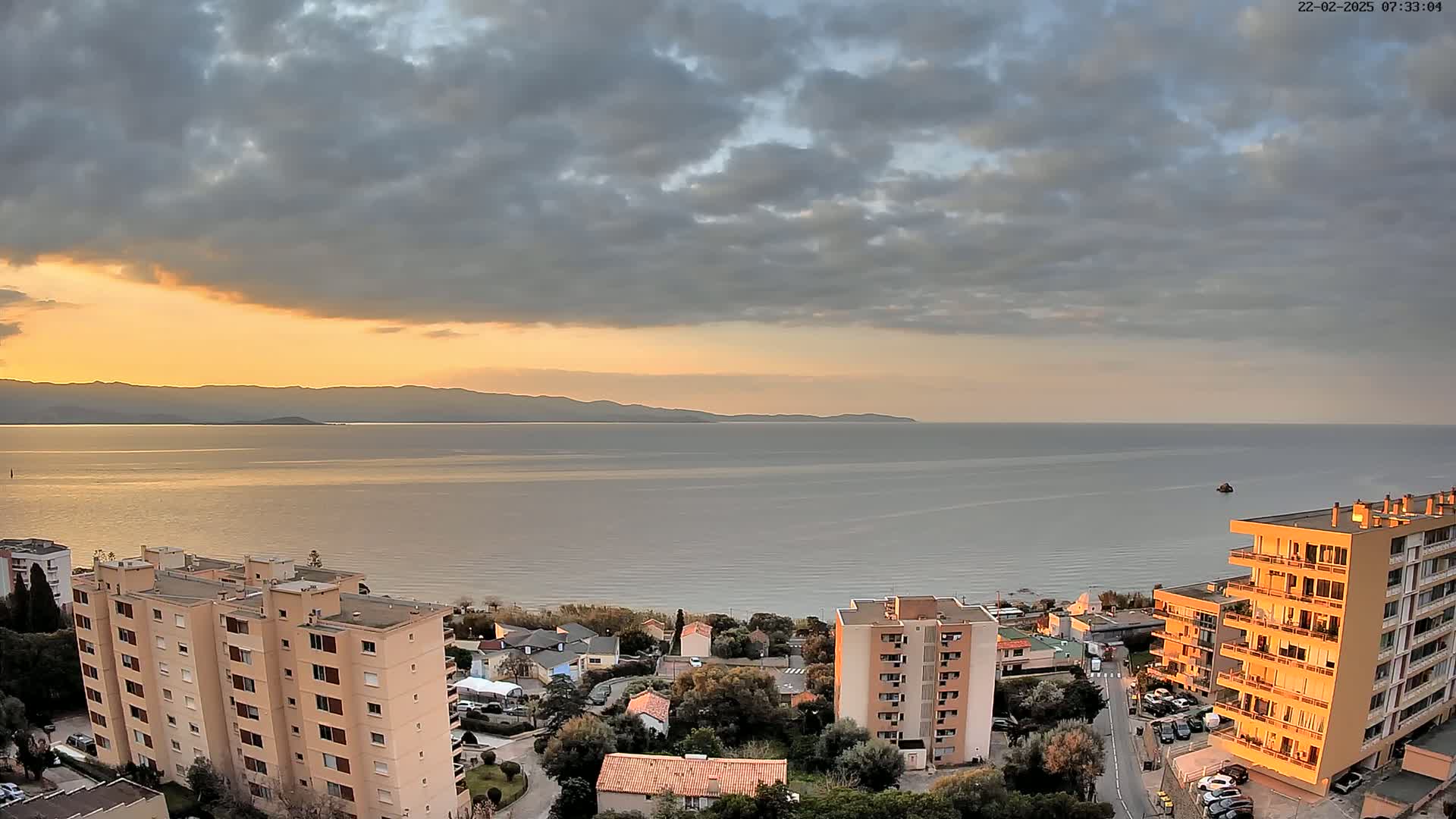 An elevated view shows a calm sea at sunset under a partly cloudy sky, with several apartment buildings and houses visible along the shoreline.