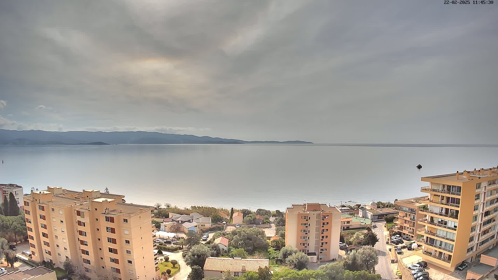A coastal scene under a cloudy sky shows a calm sea, a sandy beach, residential buildings, and distant mountains.