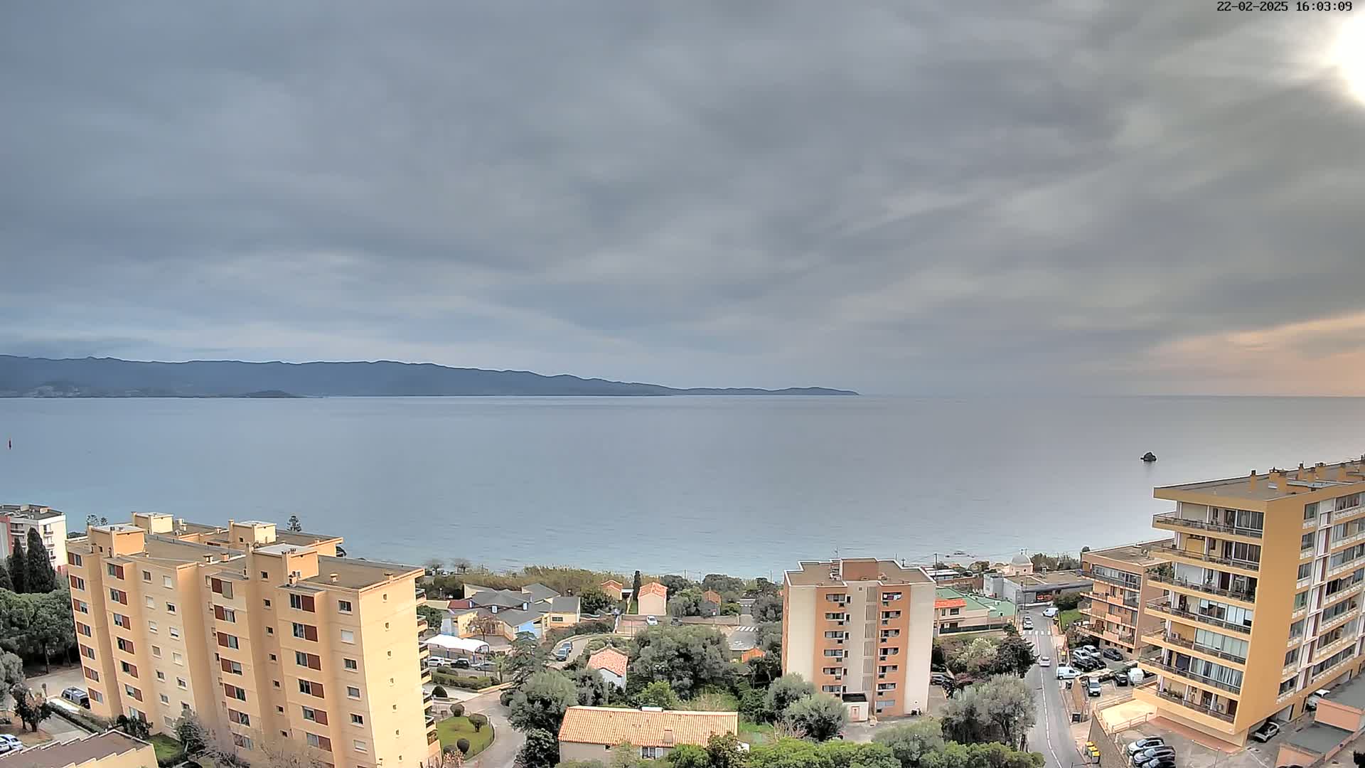 A coastal town with multi-story buildings is shown under an overcast sky, overlooking a calm sea and distant mountains.