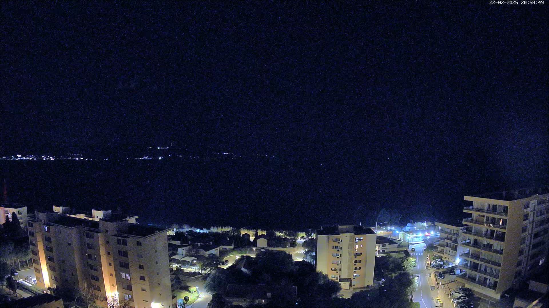A nighttime aerial view shows a coastal town with apartment buildings illuminated against a dark, clear sky and a body of water in the background.