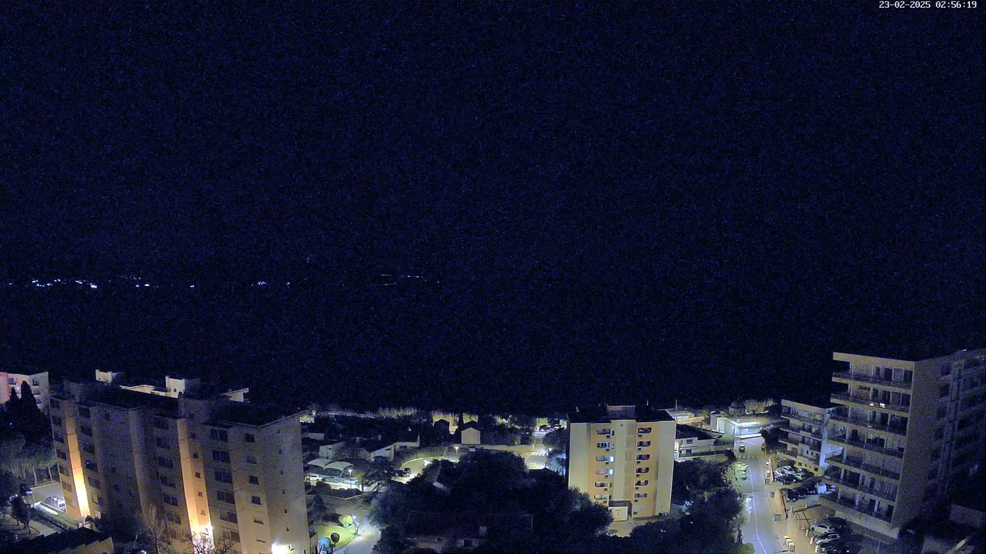 A nighttime, high-angle view shows a coastal town with several apartment buildings illuminated, and a dark body of water in the background under a clear sky.