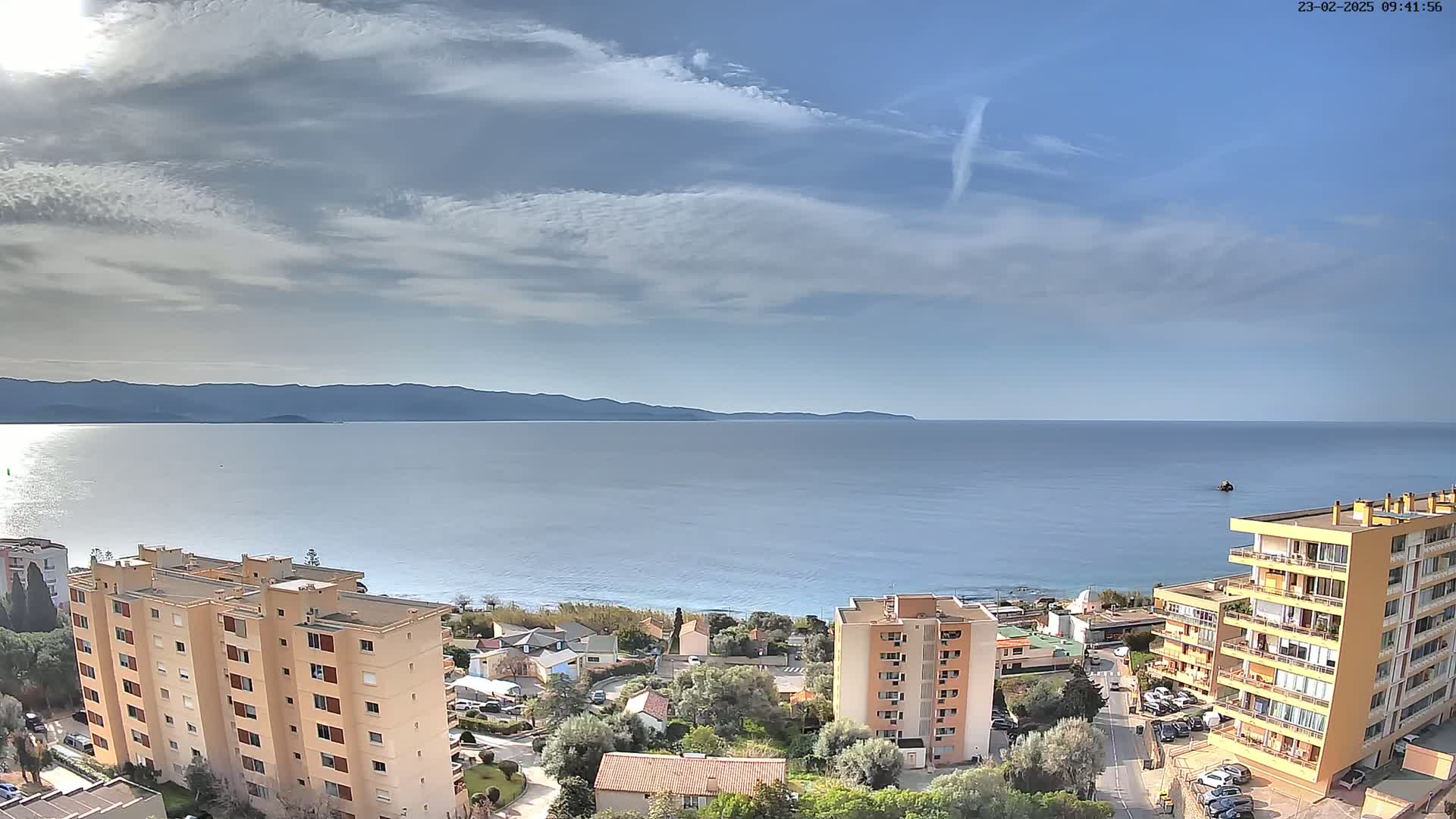 A partly cloudy day reveals a coastal scene with calm blue water, distant mountains, and several multi-story buildings nestled among low-rise structures and green vegetation.