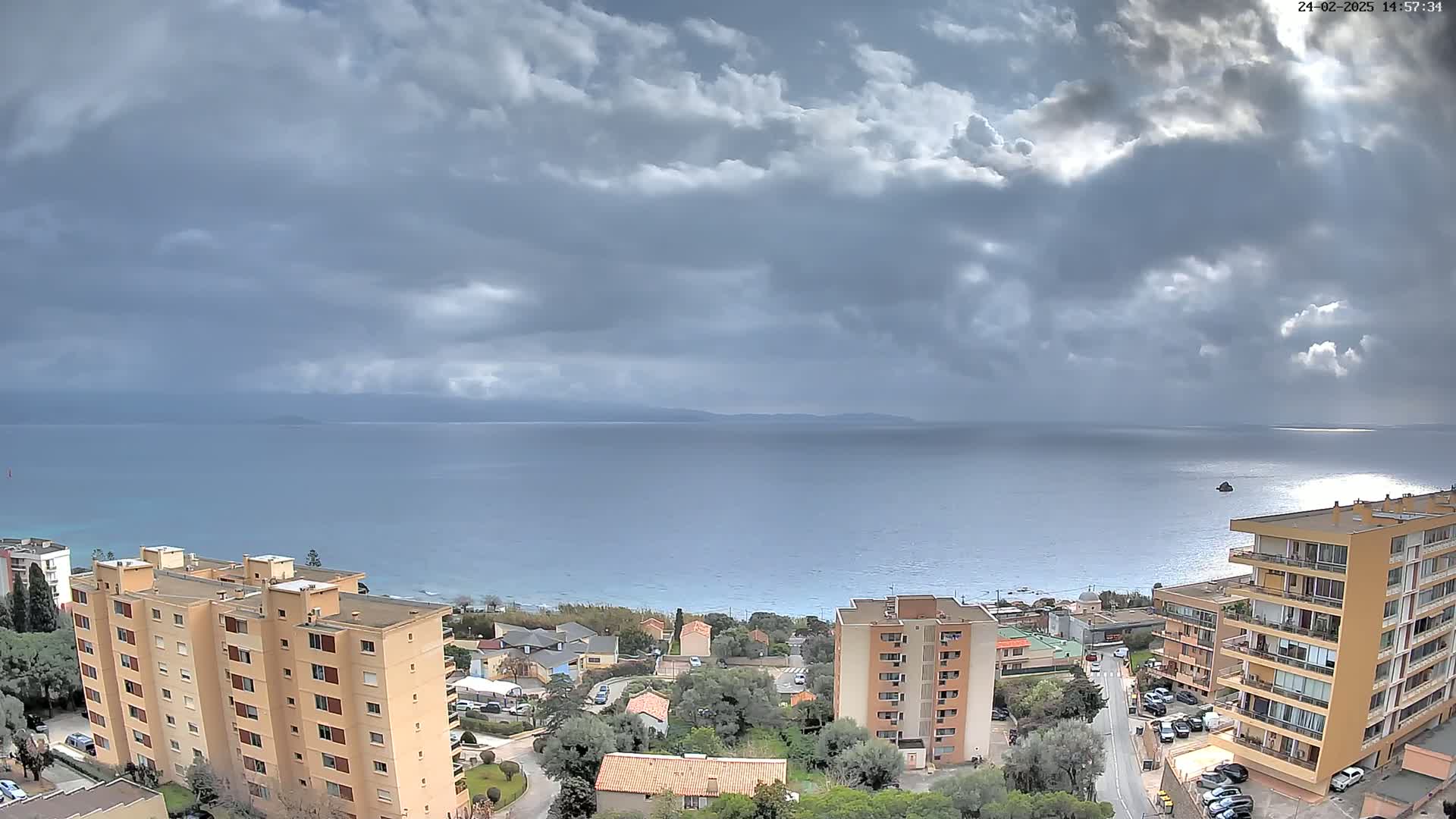 A coastal town under a partly cloudy sky overlooks a calm sea with distant mountains.