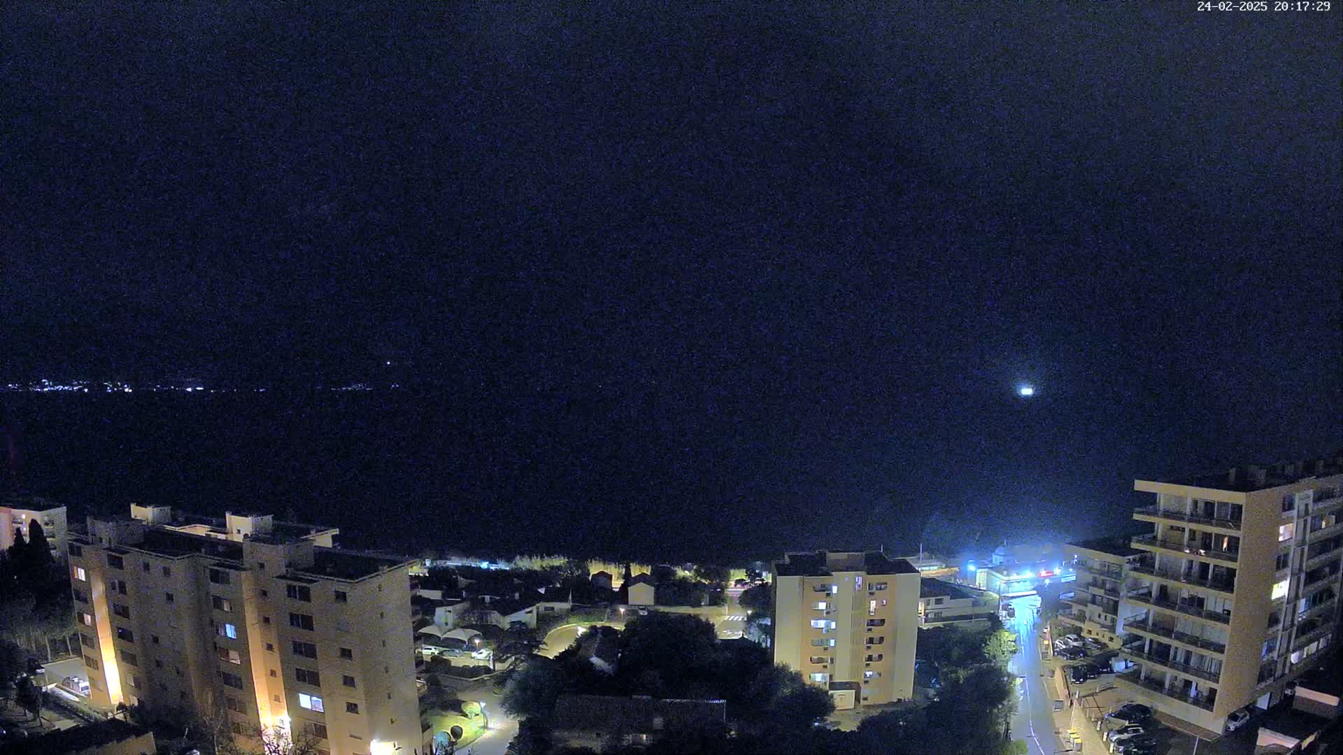 A nighttime view shows a dark body of water with a distant shoreline of lights under a clear, dark sky, overlooking several illuminated apartment buildings and a street.
