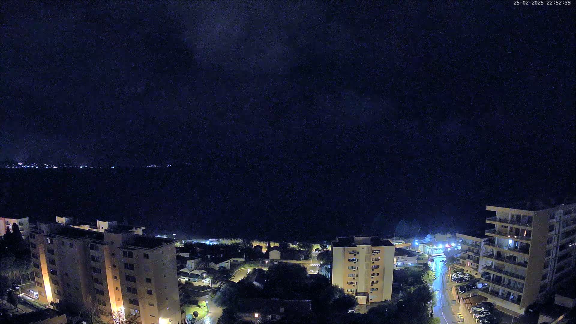 A nighttime aerial view of a coastal town with apartment buildings and a calm, dark body of water in the background under a clear, dark sky.