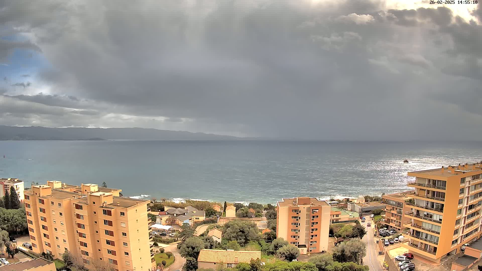 An overcast sky hangs over a choppy sea, with several apartment buildings and houses visible along the coast.