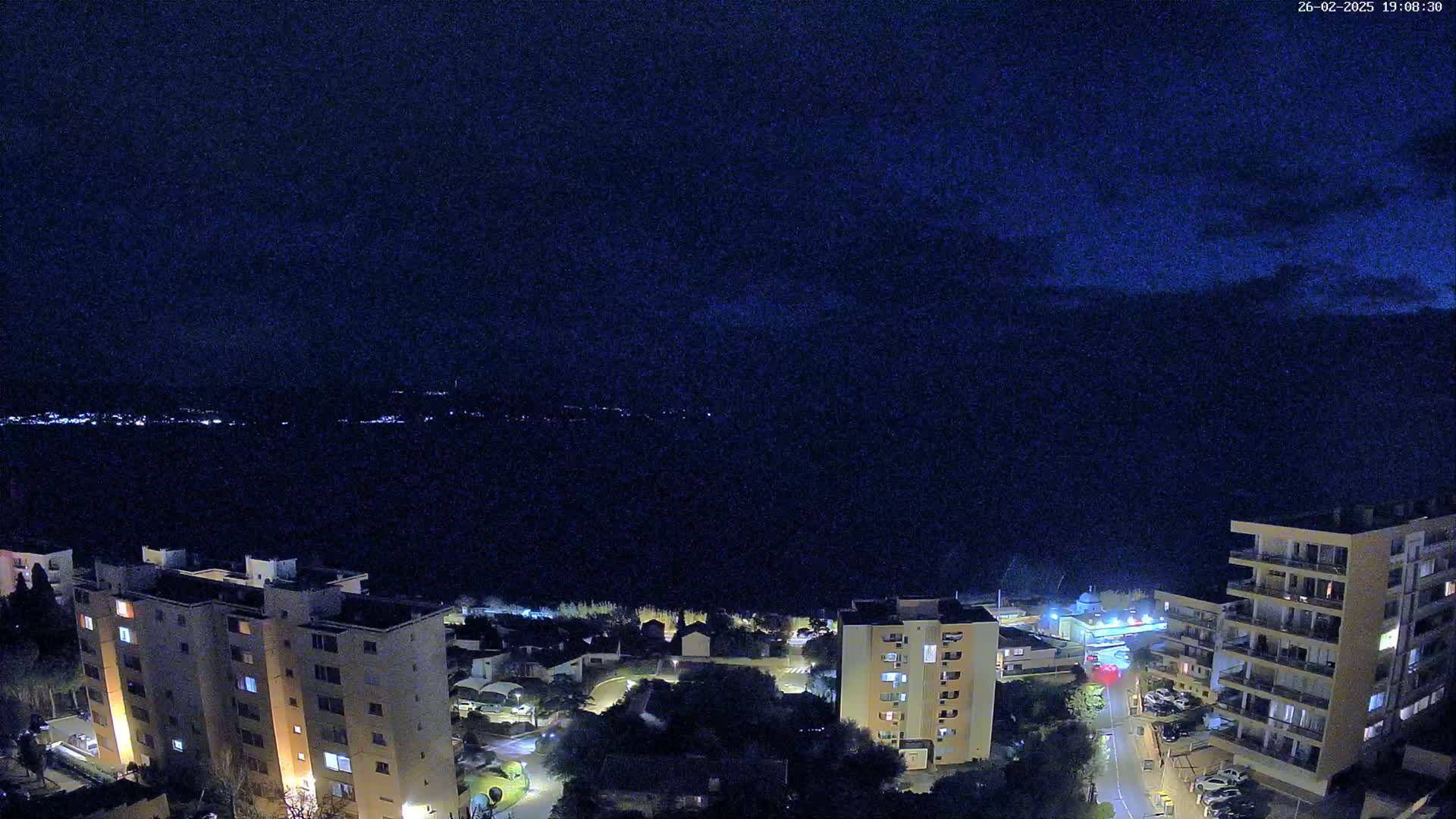 A nighttime aerial view shows a dark body of water, a cityscape with several multi-story buildings, and a mostly clear sky.