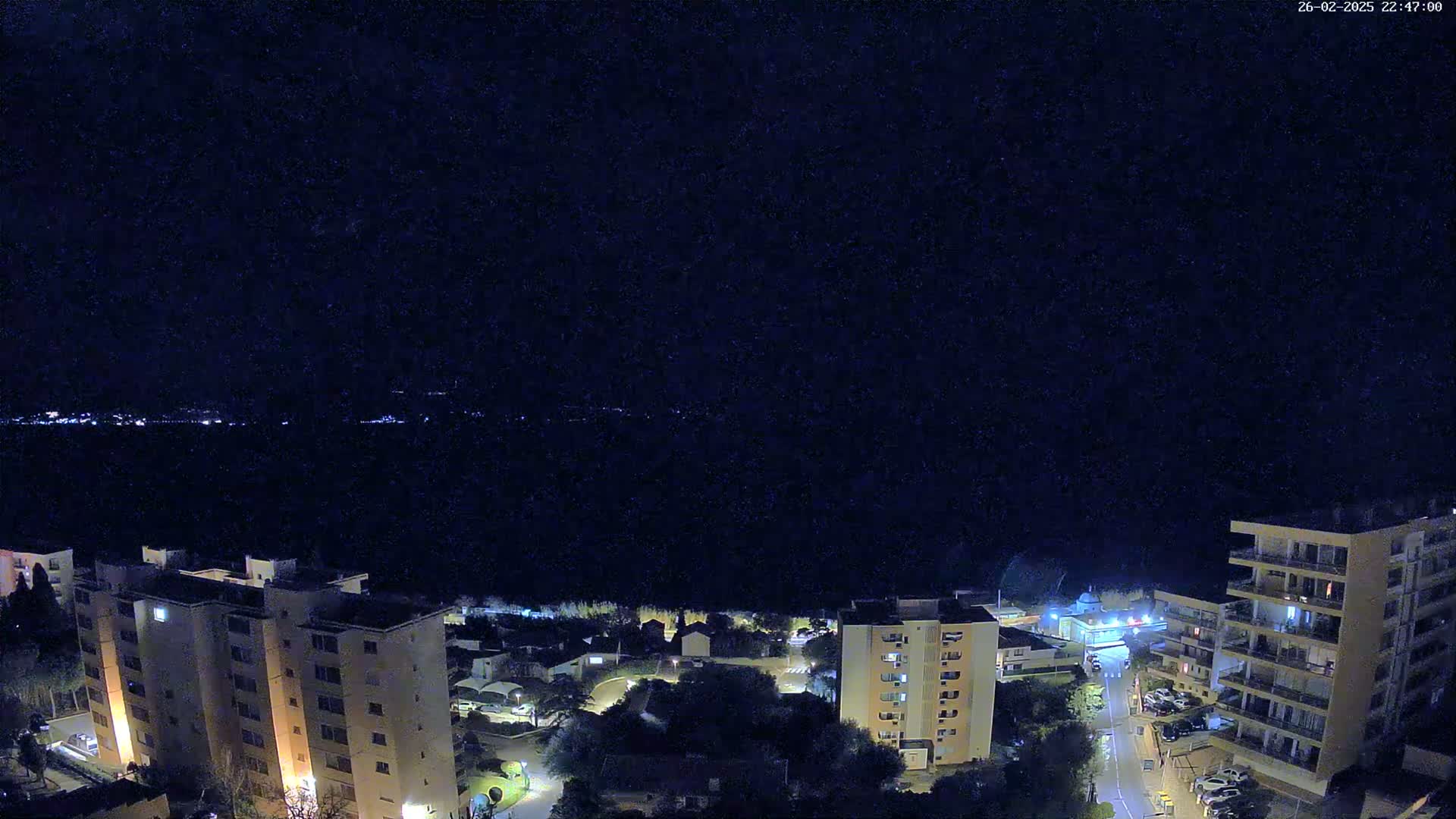 A nighttime aerial view shows a coastal town with several multi-story buildings under a clear, dark sky.