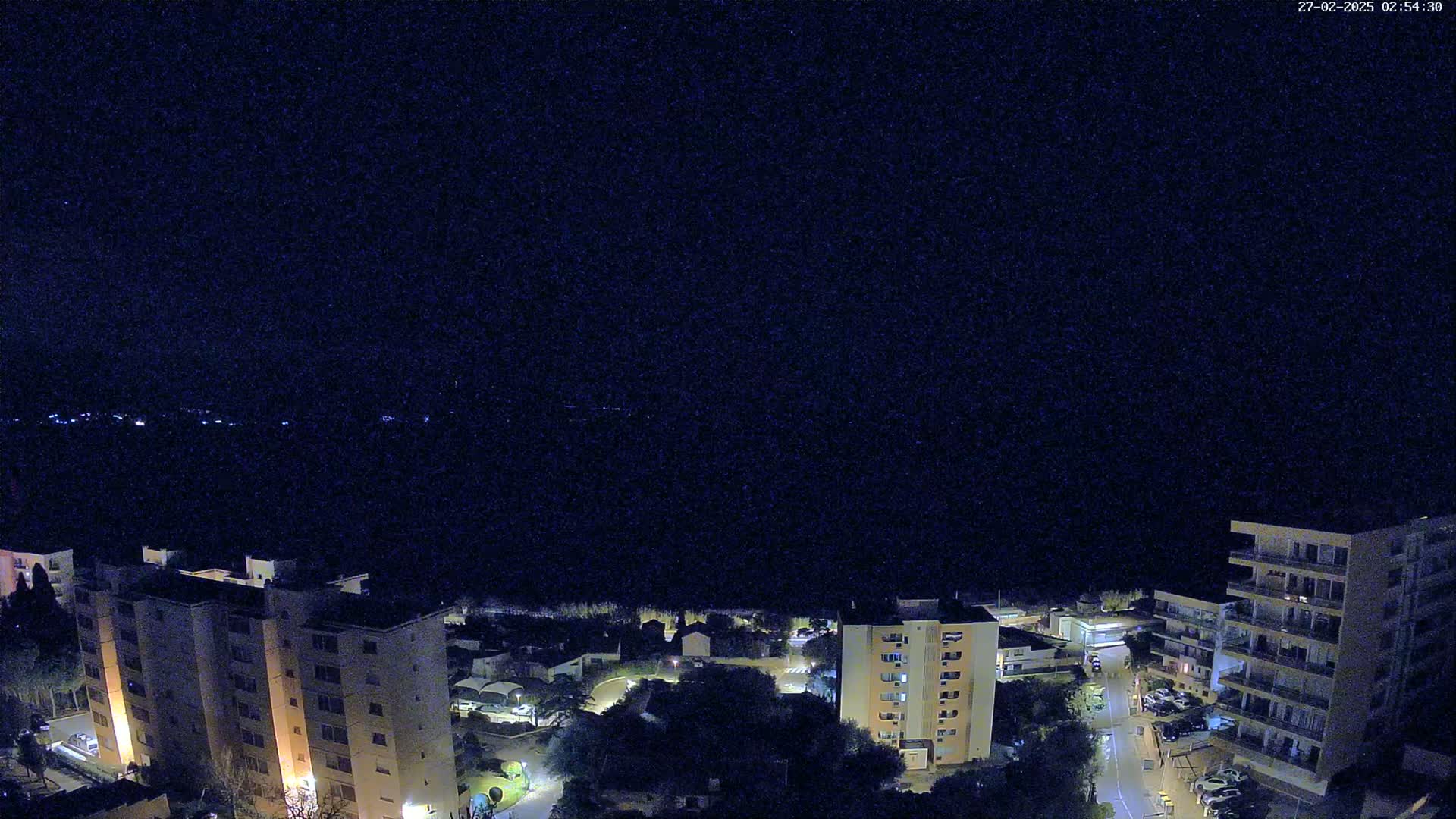 A nighttime, overhead view of several illuminated apartment buildings and surrounding streets nestled amongst dark trees under a clear, starry sky.