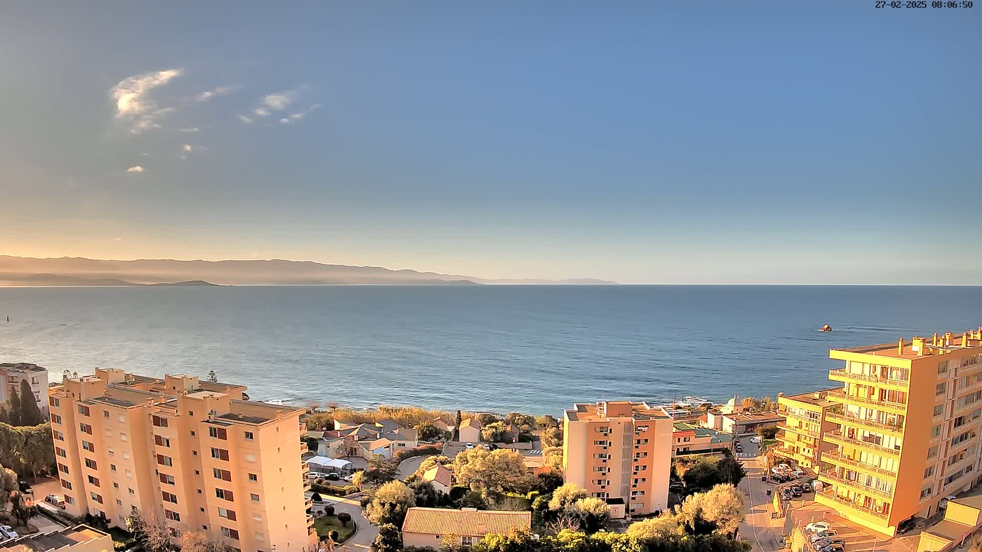 A coastal town with several apartment buildings overlooks a calm sea under a mostly clear, sunny sky, with mountains visible in the distance.