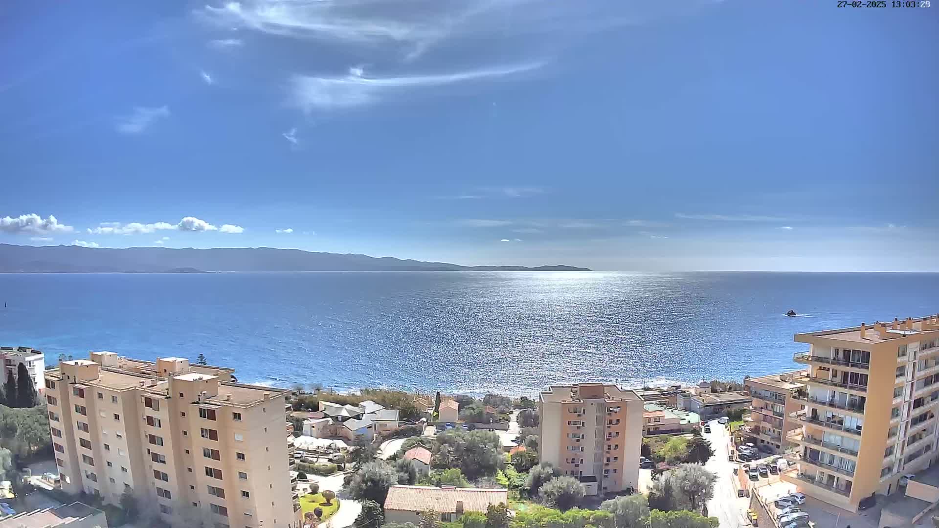 A sunny day overlooking a calm blue sea with distant mountains,  a coastal town with several apartment buildings visible in the foreground.