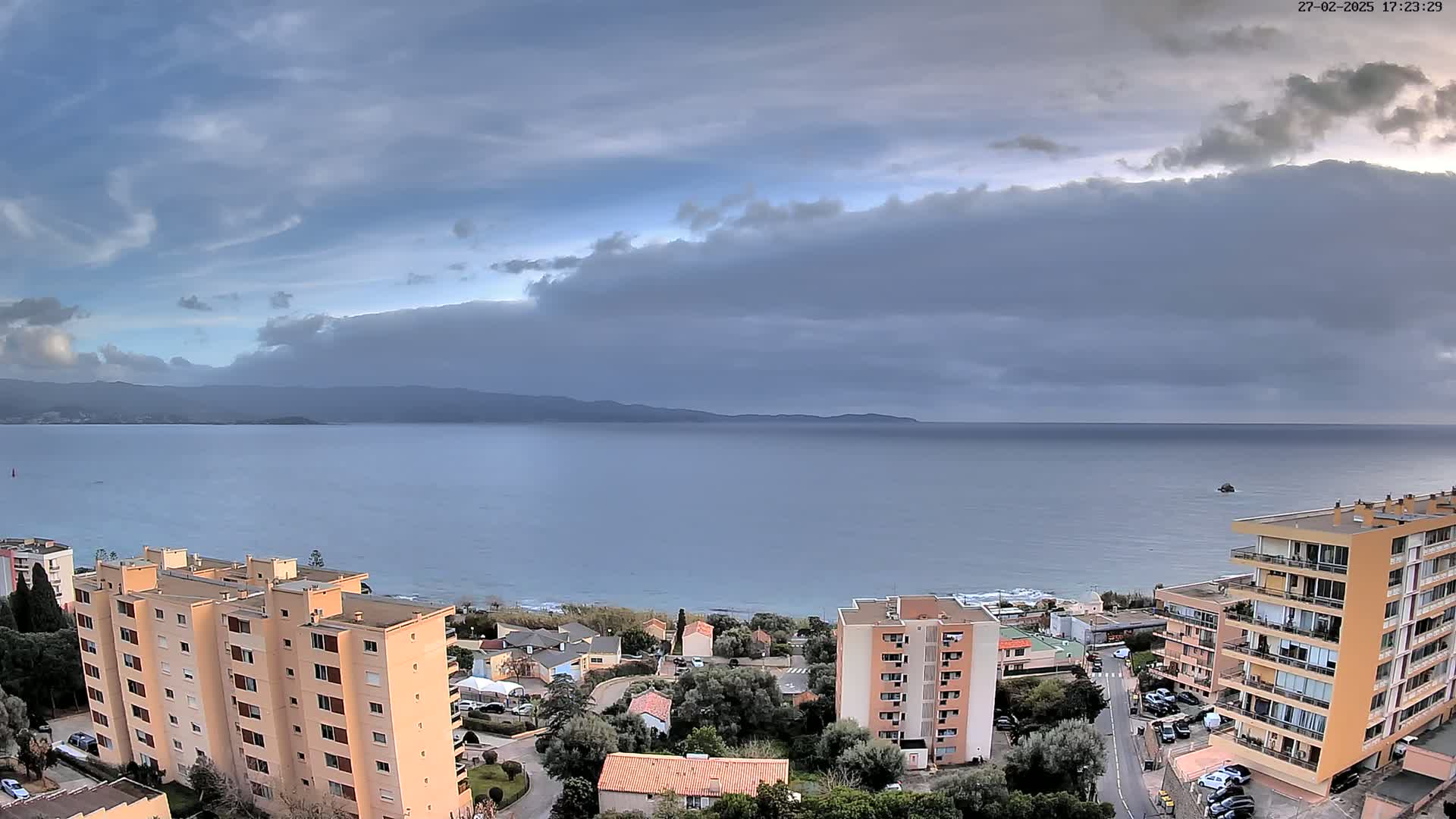 A wide, high-angle view shows a calm sea under a mostly cloudy sky, with several apartment buildings and residential areas visible along the shoreline.