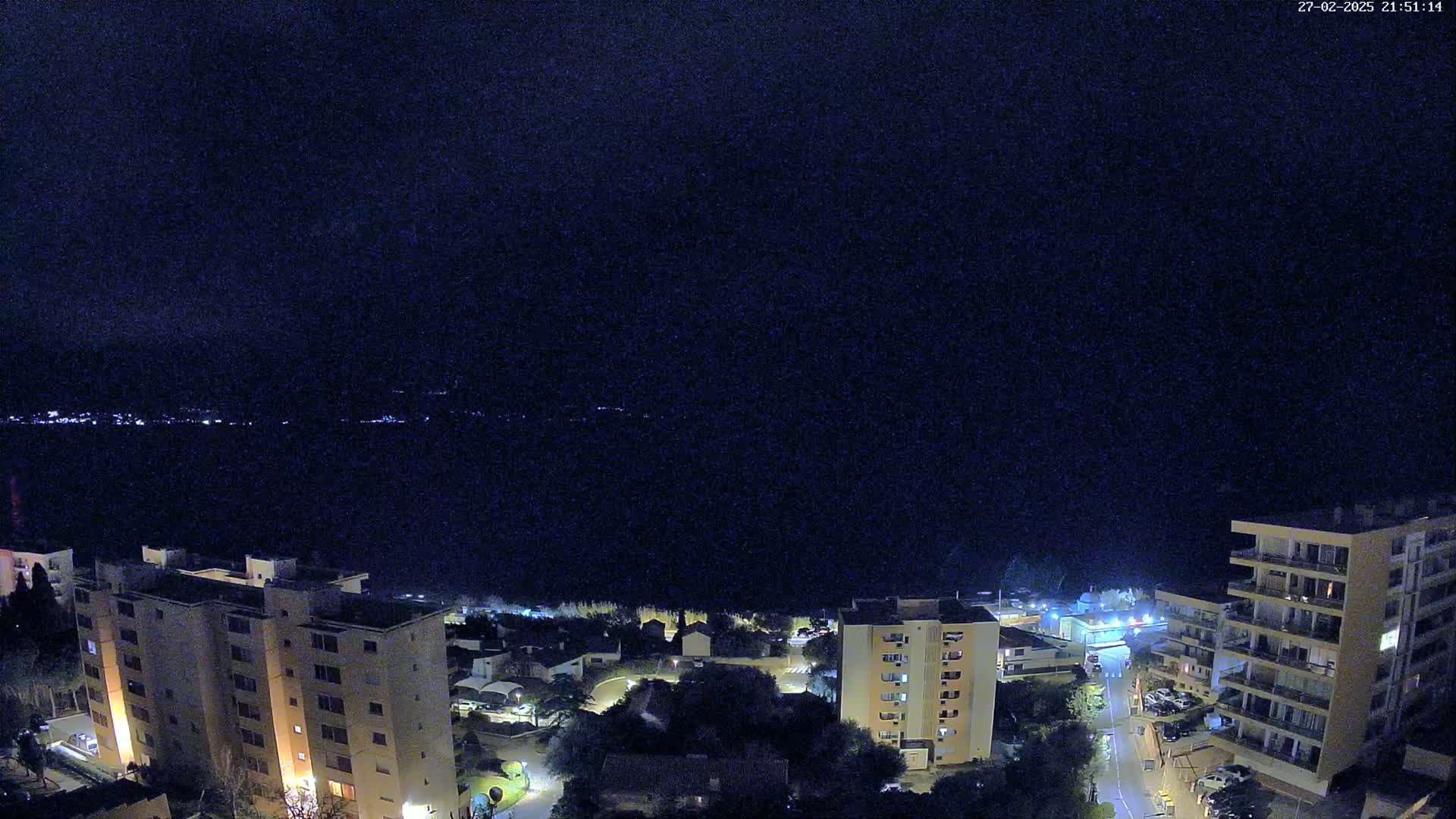 A nighttime, high-angle view of a coastal town with apartment buildings illuminated against a dark body of water under a clear, dark sky.