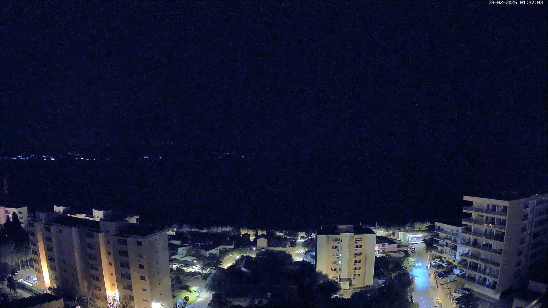A nighttime aerial view shows several apartment buildings with lights on, a road with a few cars, and a dark, clear sky.