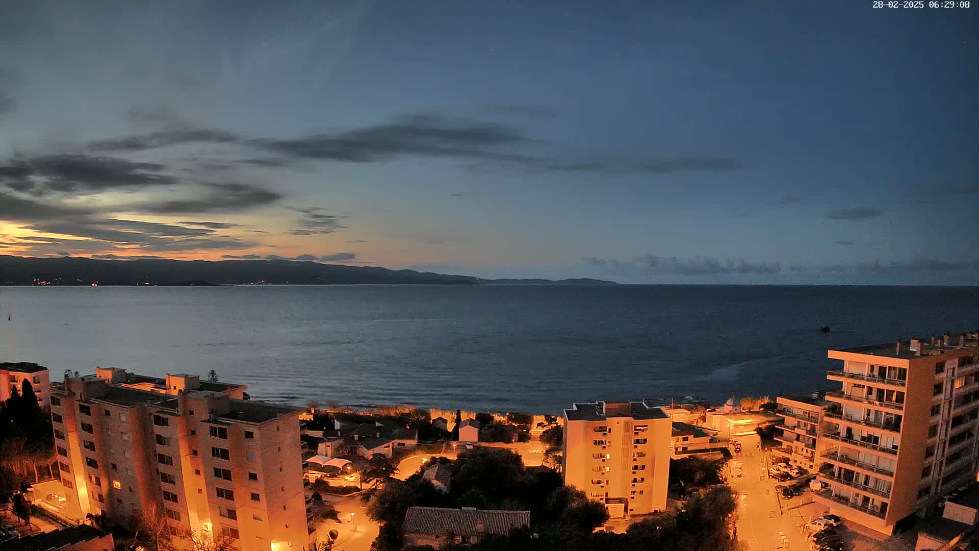 A coastal town at twilight, with calm water and dimly lit buildings under a partly cloudy sky.