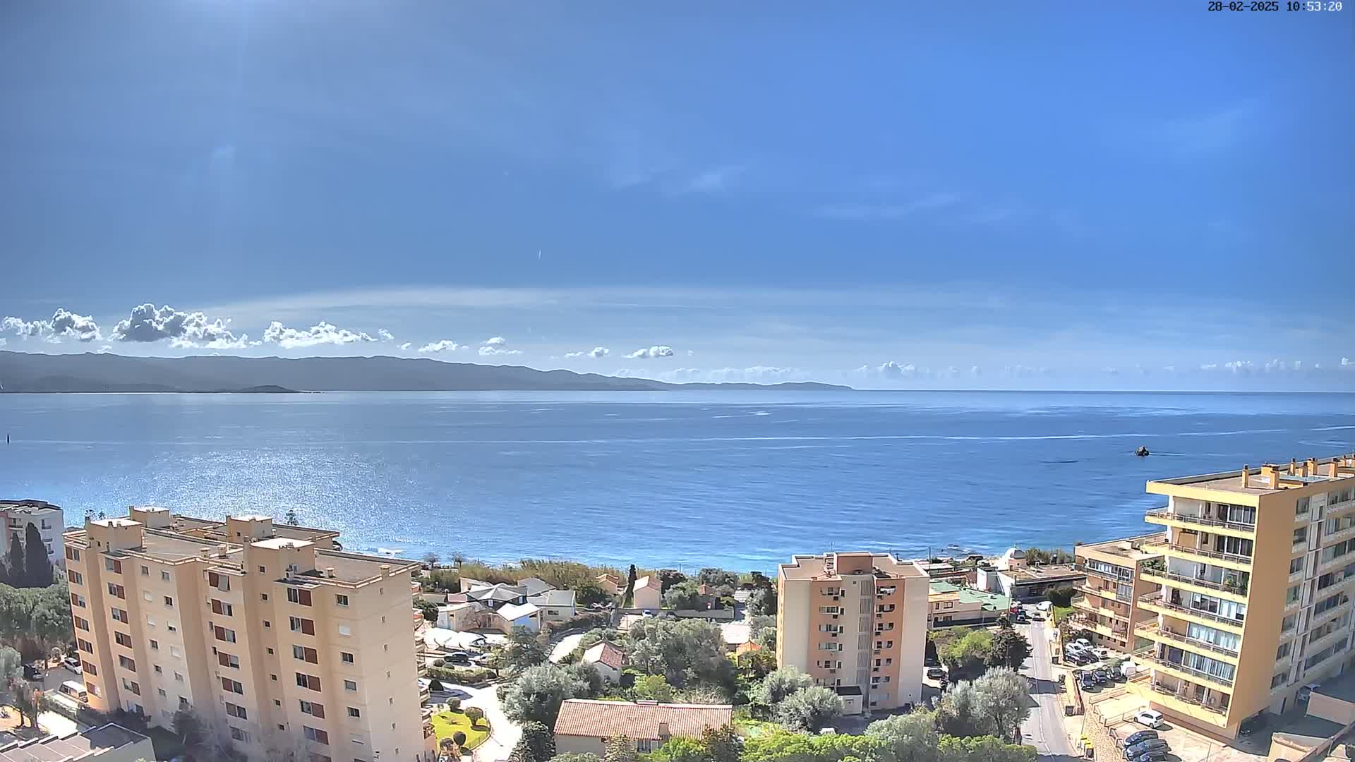 A sunny day reveals a coastal scene with apartment buildings overlooking a calm sea and distant mountains under a mostly clear sky.