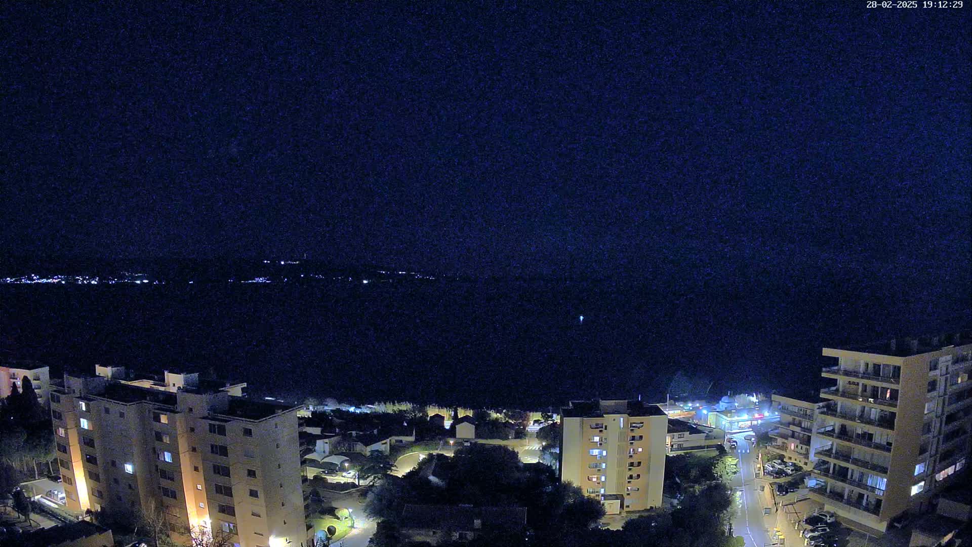 A nighttime aerial view shows a dark body of water with distant city lights, and a coastal town with several apartment buildings illuminated.