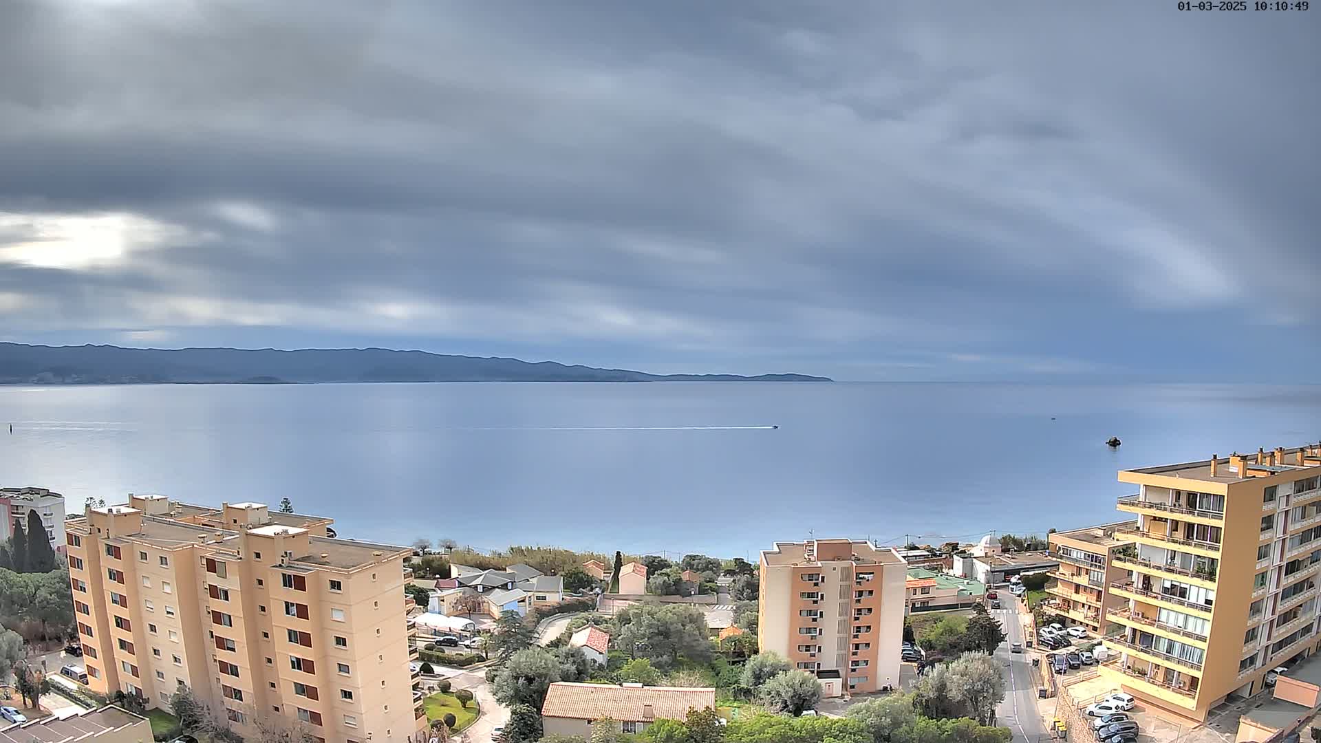 A mostly cloudy day overlooks a calm sea with distant mountains, apartment buildings, and houses clustered near a shoreline.